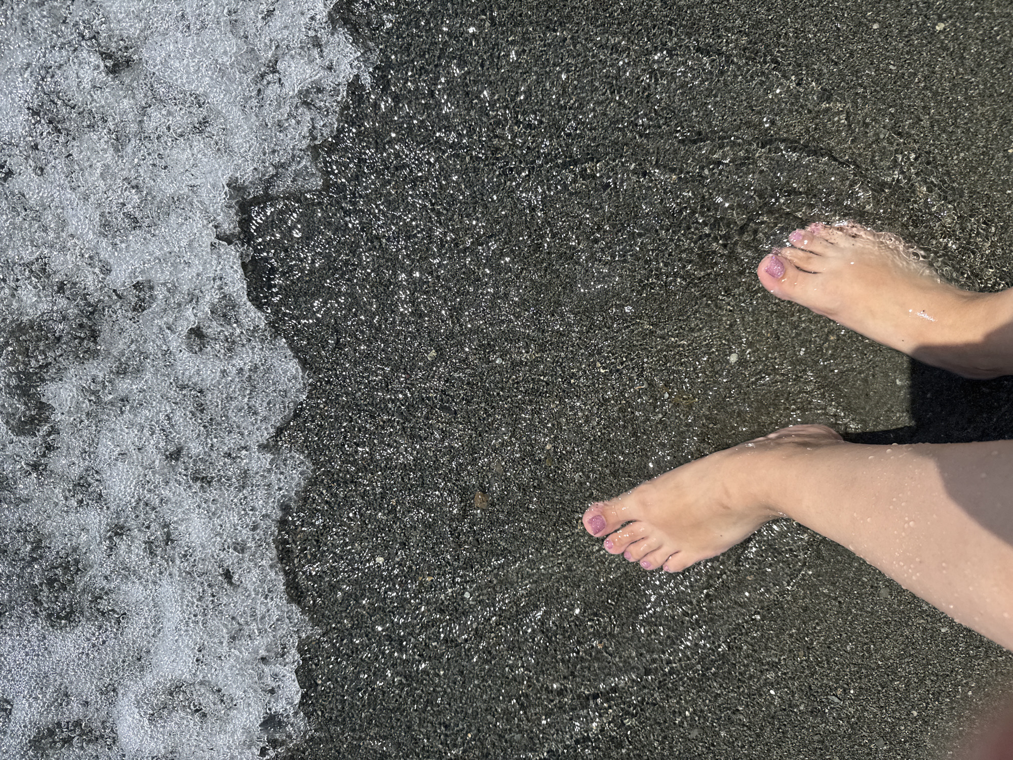 In Tsamakaberd, Armenia, the image captures a close-up, overhead view of two bare feet partially submerged in shallow, clear water on a dark, pebble-strewn beach. The water is frothy and effervescent along the edge, indicating a gentle wave. The sand or pebbles are dark gray and glisten with moisture, reflecting sunlight. The feet, with pink-painted toenails, are positioned with the toes of the right foot pointing towards the upper right and the left foot angled slightly to the left. Water droplets are visible on the skin of the legs and feet, suggesting recent immersion. The lighting indicates a bright, sunny day.