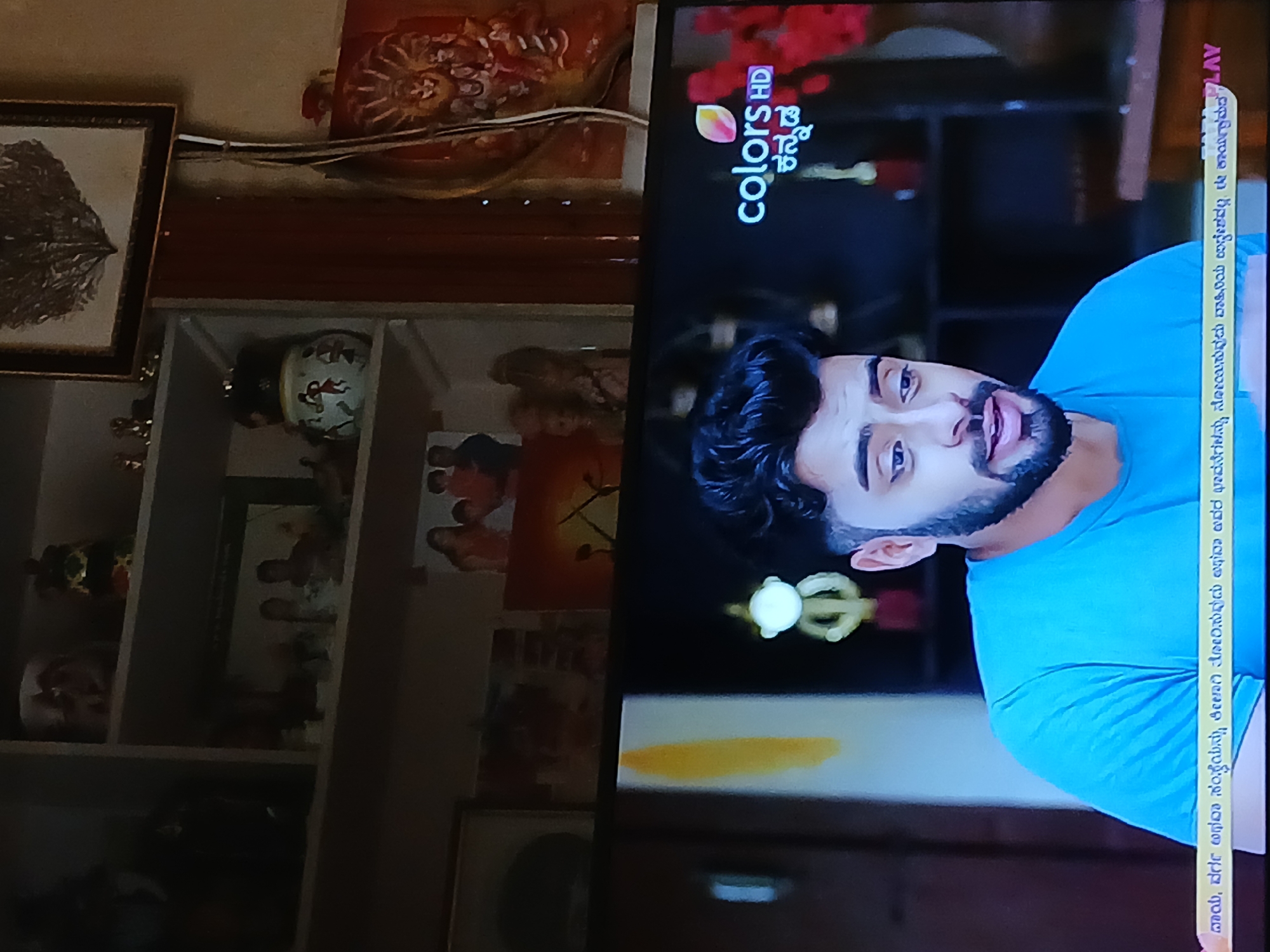 Here is a detailed description of the image:
**Scene:** The image shows a close-up shot of a television screen displaying a scene from the "Colors Kannada" television channel.
**Subjects:**
* A male actor is visible on the screen, wearing a light blue T-shirt. He has dark hair and a beard.
**Objects:**
* The television screen displays the "Colors Kannada" logo and a promotional message in Kannada.
* Behind the television, there's a shelf with various decorative items:
* A framed picture.
* Decorative vases.
* Smaller frames and other small objects.
* There is a small, decorative item above the shelf, which looks like a statue.
**Environment:**
* The scene takes place indoors.
* The background has some items that appear to be placed on the shelf, including artwork and decorative pieces.
* The lighting in the room is soft, but there are clear reflections on the television screen.
**Inferences:**
* "Colors Kannada" is a Kannada-language general entertainment channel.
* The promotional message at the side of the screen serves as a legal disclaimer.
**Location:** Although the channel is related to the Kannada language and culture, which is primarily associated with the state of Karnataka in India, there is nothing in the image to identify the location specifically as Bengaluru or any other city in India.