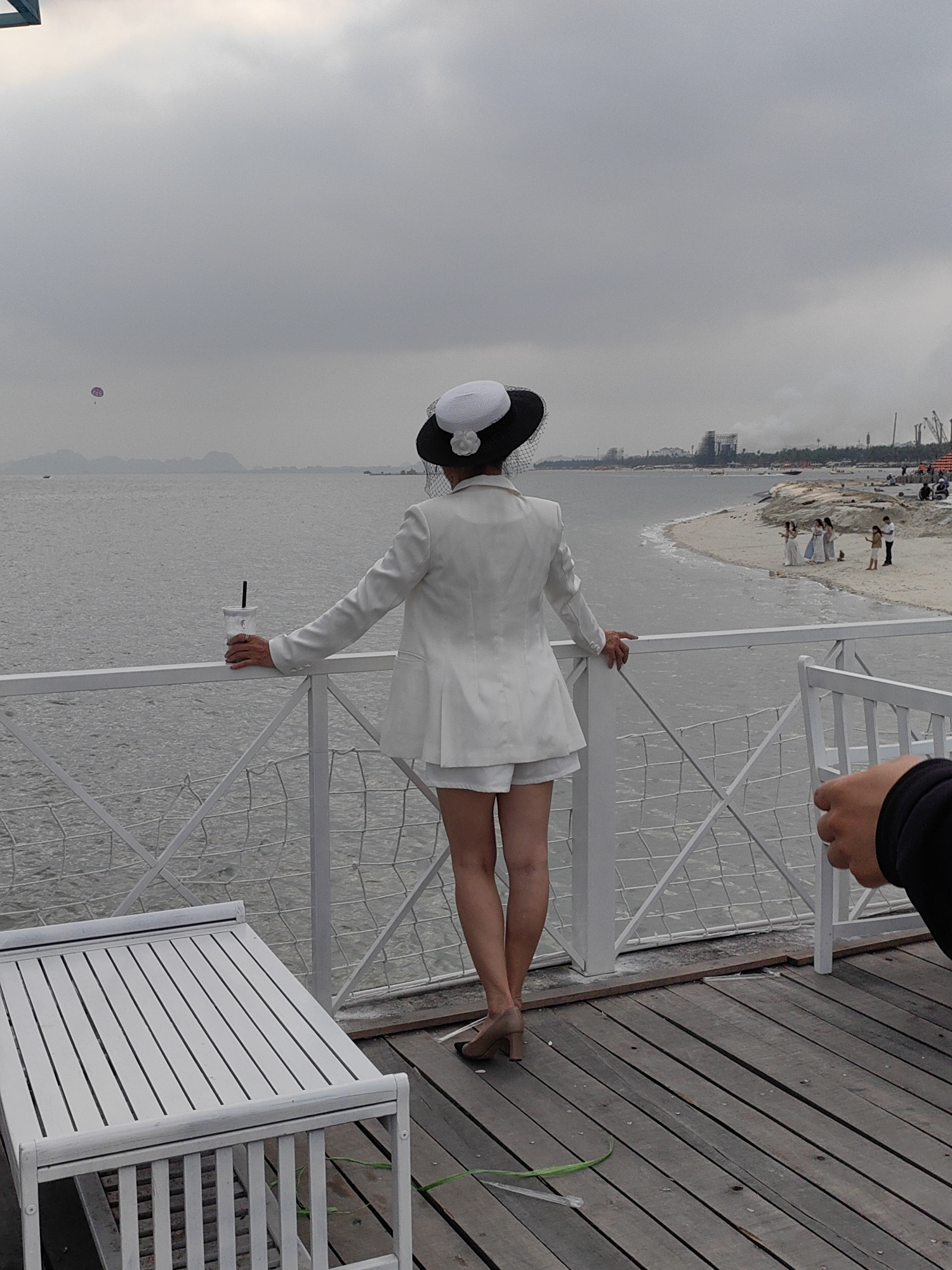 A woman, viewed from behind, stands on a wooden deck, leaning against a white railing and gazing out over a body of water. She is dressed in a white blazer, white shorts, and light brown block-heeled shoes. A distinctive black and white hat with a white flower and a fine net veil adorns her head. Her right hand holds a clear plastic cup with a dark straw, resting on the railing, while her left hand also holds the railing.
The deck is made of dark wooden planks, and a white slatted bench or table is positioned in the lower-left foreground. The railing features a crisscross mesh design in its lower section.
Beyond the railing, a calm, grey body of water extends to the horizon. In the distance, a sandy beach curves along the coastline to the right. Several people are visible on the beach; some appear to be posing for photos, dressed in light-colored gowns, possibly indicating a wedding event or photoshoot. Further down the beach, construction equipment, including cranes, is discernible, suggesting ongoing development.
In the far distance over the water, a lone individual is parasailing with a small pink parachute. Hazy hills or mountains line the distant horizon, and a small boat is also present on the water. The sky is uniformly overcast and grey, creating a subdued, cloudy atmosphere, typical of daytime on a non-sunny day. Part of another person's arm and hand, wearing a dark sleeve, is visible in the lower right foreground. No visible text is present in the image. The scene is consistent with a coastal area like Bai Chay Ward, Vietnam.