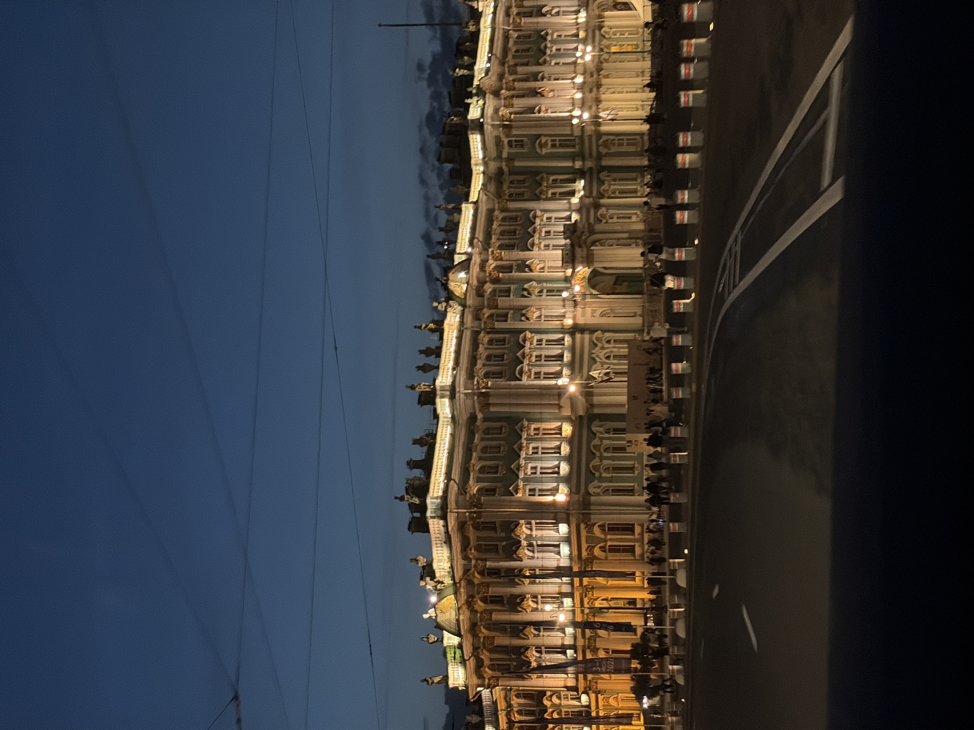 This image shows a nighttime view of the State Hermitage Museum, formerly the Winter Palace, in St. Petersburg, Russia. The building is illuminated with warm lighting, highlighting its ornate architecture, including columns, statues, and numerous windows. A dark road with white lane markings is in the foreground, with a line of white and red traffic barriers separating it from the museum's plaza. A crowd of people, mostly silhouetted against the building's light, are gathered on the plaza. The sky above is a deep twilight blue with some subtle cloud formations. Overhead wires are visible against the sky.