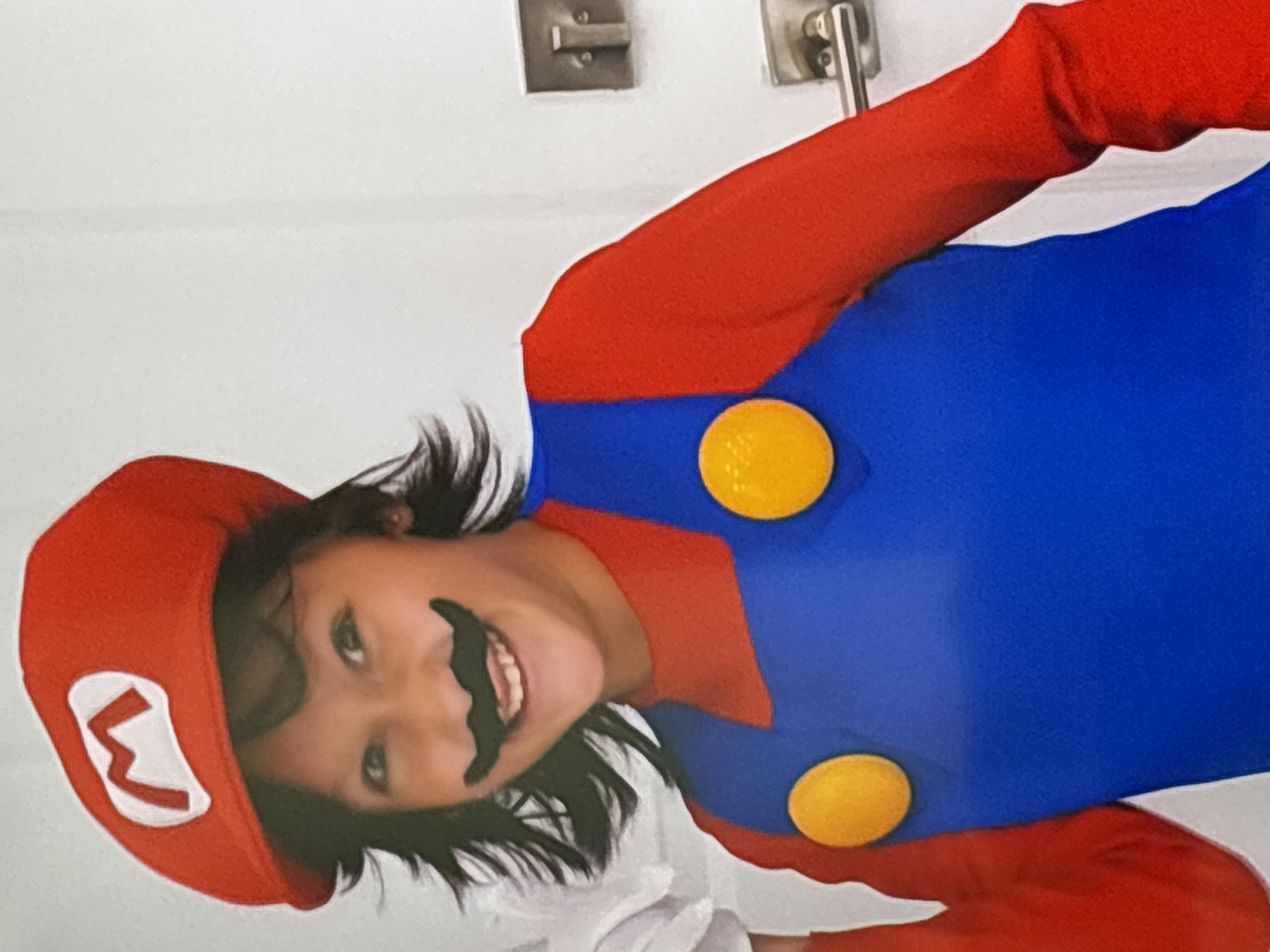 A young child wearing a red cap with a white and red "M" logo, a fake black mustache, and a red and blue Mario-themed costume smiles at the camera. The child's dark hair is visible around the cap. The background is a plain white wall with two silver door handles. The image appears to be taken indoors, possibly in a home. There is no indication of this scene being in Dubai, United Arab Emirates.