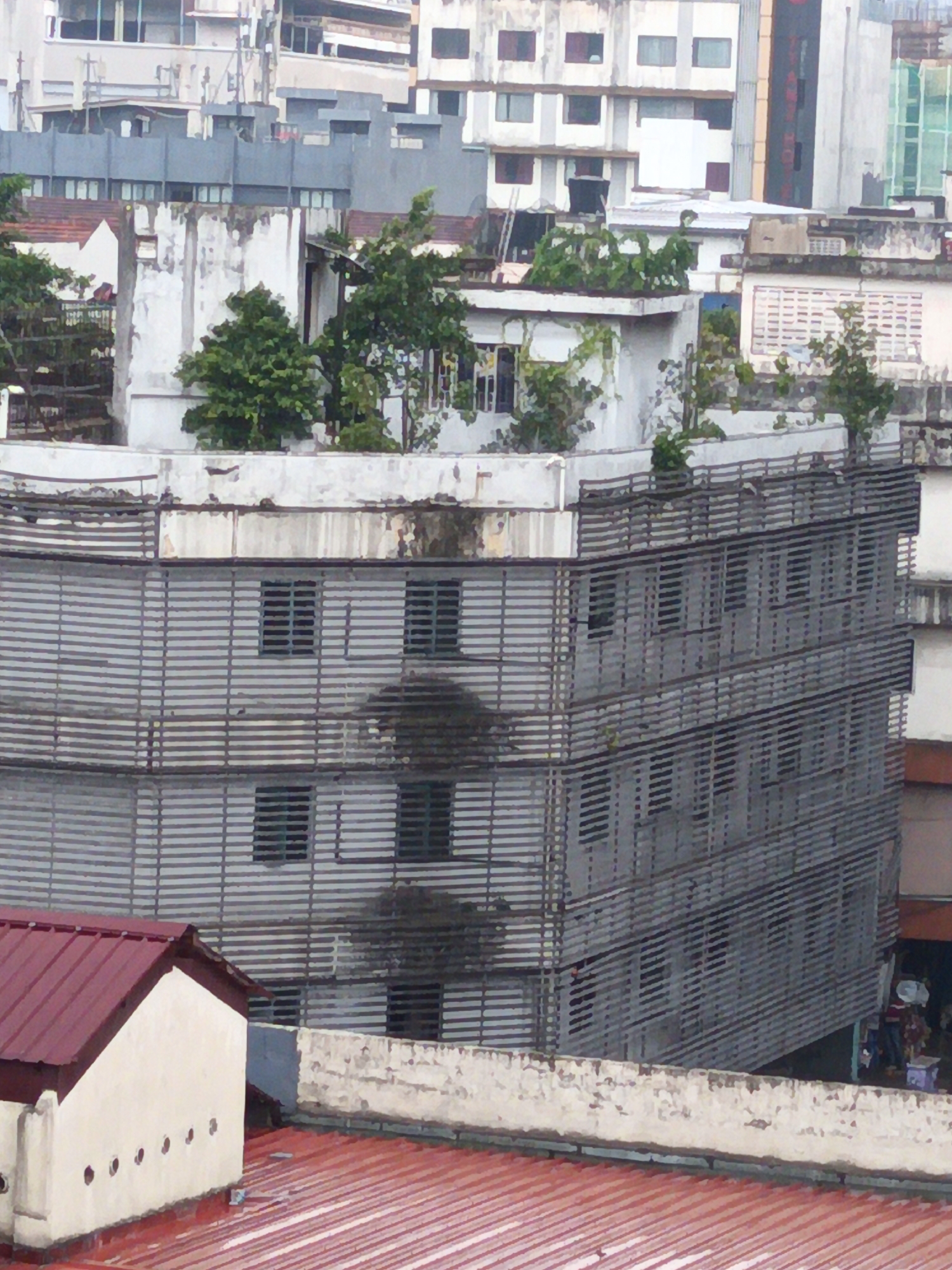 This is an aerial shot of a densely built urban environment, likely in Kuala Lumpur, Malaysia, given its architectural style and the presence of tropical foliage. The image is dominated by a multi-story building with a distinctive horizontal louvered facade, suggesting a humid climate where such features are common for ventilation and sun shading. The building appears to be constructed from concrete and metal.
In the foreground, a building with a red, sloped roof made of corrugated metal is visible. It has a cream-colored wall with a series of circular openings. Adjacent to it is another section of red corrugated roofing. The overall impression is of a mix of older and more modern construction, typical of many Southeast Asian cities.
Several trees with dense green foliage are interspersed amongst the buildings, indicating that the area is not completely devoid of vegetation. The sky is not visible in this frame, but the lighting suggests it is daytime. The scene has a slightly hazy quality, possibly due to atmospheric conditions or the image processing. No people are clearly discernible in the image, though there are hints of activity on the ground level of the louvered building on the right side, with what appears to be stalls or shops with colorful awnings and figures in the distance. The composition focuses on the architecture and the urban landscape.