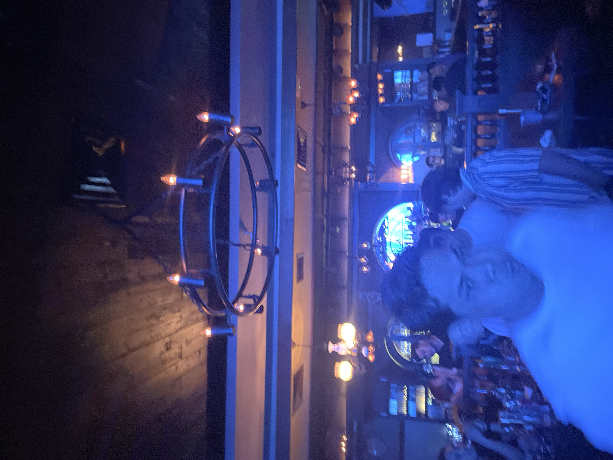 The image depicts a dimly lit interior of what appears to be a bar or restaurant. The lighting is predominantly blue, with some amber accents from the light fixtures. In the foreground, a person wearing a white shirt is facing the camera with a serious expression. Behind this person, other individuals are visible, some appearing to be patrons and others possibly staff.
The environment features wooden elements on the ceiling and decorative light fixtures, including a large, circular chandelier with candle-like bulbs. In the background, a bar area is discernible with shelves of bottles and glassware. The overall ambiance suggests a social gathering or a night out. The location is identified as Central Jakarta, Indonesia.