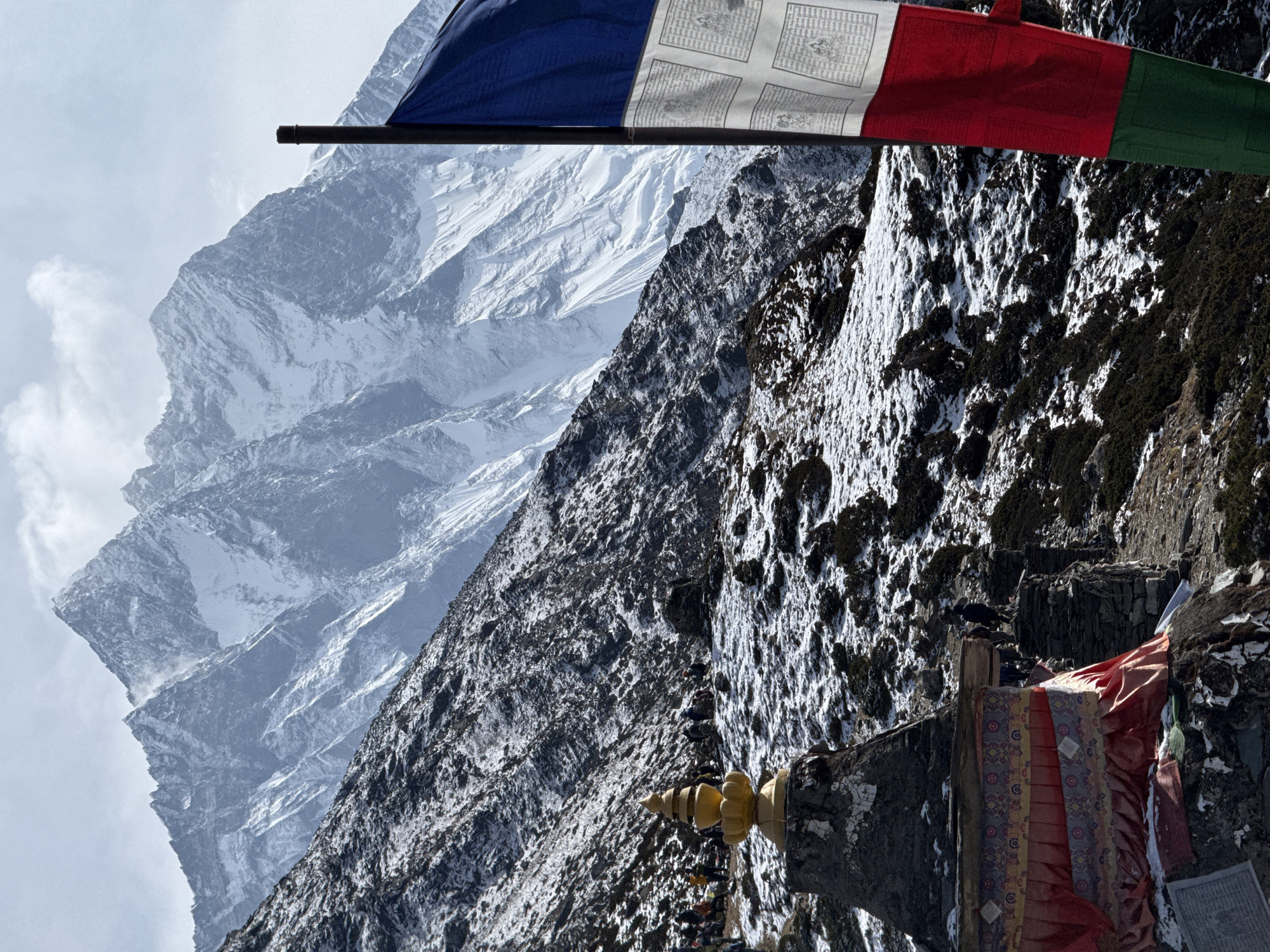 The image depicts a mountain landscape in Khumjung, Nepal, possibly taken from a high altitude. Dominating the scene are two distinct mountain slopes: one covered in snow and the other interspersed with dark vegetation and patches of snow. A string of prayer flags in blue, white, red, and green colors is positioned at the top of the frame. In the lower right corner, there appears to be a stone structure with what seems to be a golden spire and draped fabric. The image has a strong emphasis on the mountainous terrain and its unique characteristics.