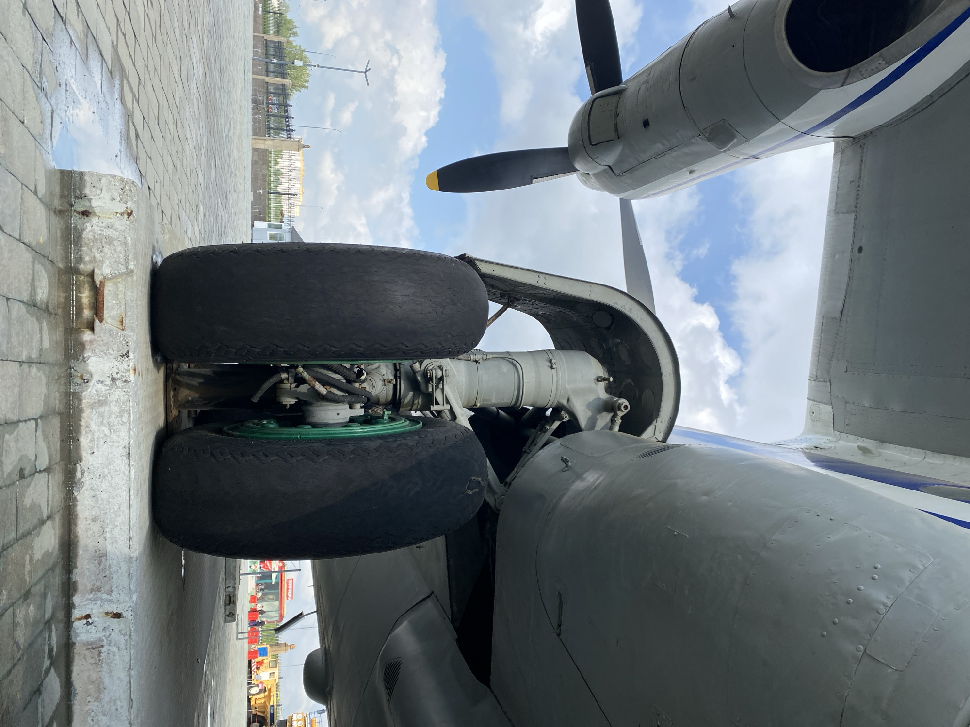 An exterior, low-angle, ground-level shot focuses on the landing gear of a large aircraft, likely an Antonov An-24, parked on a paved surface. The aircraft's massive tires, with green rims, dominate the foreground, resting on a concrete block. The underside of the plane, with its complex mechanical structure, is visible, along with a propeller in the background. The sky is partly cloudy with a bright, overcast daylight. In the distance, a fence, lampposts, and a yellow industrial vehicle can be seen, suggesting an outdoor, possibly museum or airfield, location. The scene is located in the city of Verkhnyaya Pyshma, Russia, specifically within the Military Technology Museum of UMMC, where such aircraft are often displayed. No people are visible in the frame.
