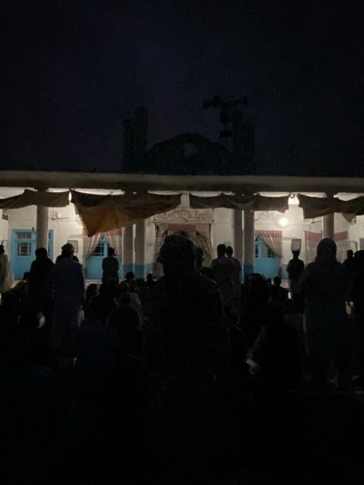 A low-angle, long-shot shows a mosque in Kandahar, Afghanistan at night with an unlit courtyard filled with dark silhouettes of many people, some sitting and some standing. The architectural features of the mosque include a building with white columns supporting a roofed area that's lit from the inside. Drapes or cloth hang down from the roof. Behind the covered area is the mosque itself, which has a central arch and twin minarets. A speaker system is positioned atop the minarets. The sky is dark, suggesting either night or a very overcast day. Details are obscured due to the dark conditions and possible low lighting.