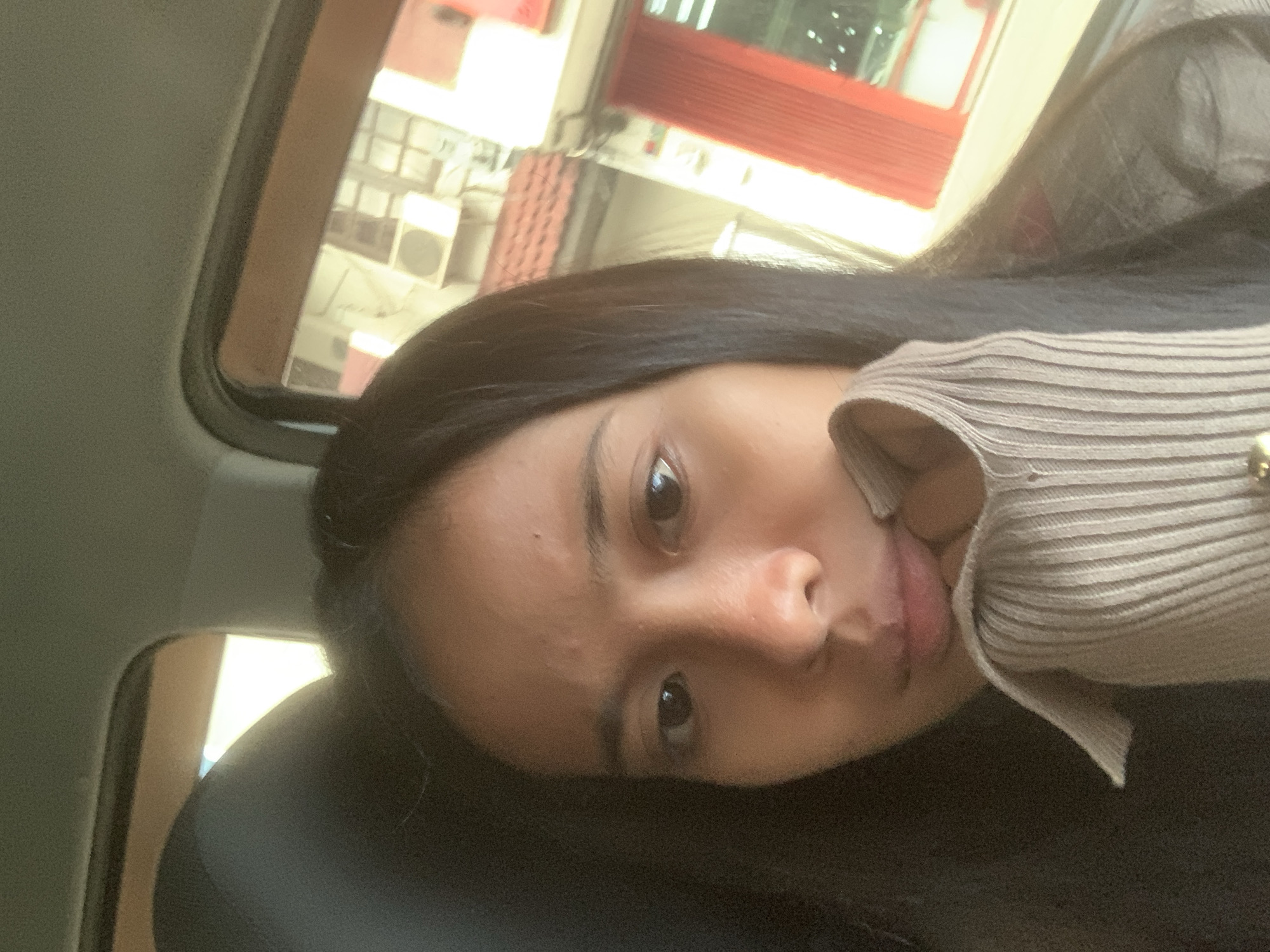 A person with dark hair and brown eyes is lying down, possibly in a car, as a car window frame is visible. The background includes a blurred view of buildings, one with a red exterior, suggesting an urban area. The picture might have been taken in Samarinda, Indonesia, but location information is uncertain due to the ambiguity and the blurred image of the background. The person is wearing a beige sweater, partially obscuring their face. The focal point is the individual's face.