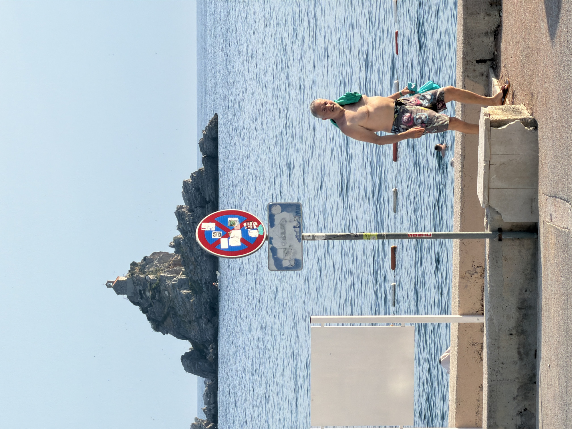 A man in swim trunks walks along a concrete embankment by the sea in Petrovac na Moru, Montenegro. He is shirtless, with a green towel draped over his left shoulder and a turquoise bag in his right hand. He is wearing flip-flops. In the background, the sea stretches to the horizon, with a rocky island topped by a small church visible in the distance. A red and blue "no parking" sign is mounted on a pole to the left of the man, partially obscuring a white rectangular board. The embankment is constructed of concrete and stone. The sky is clear and blue. Another person is visible in the water further out in the sea.