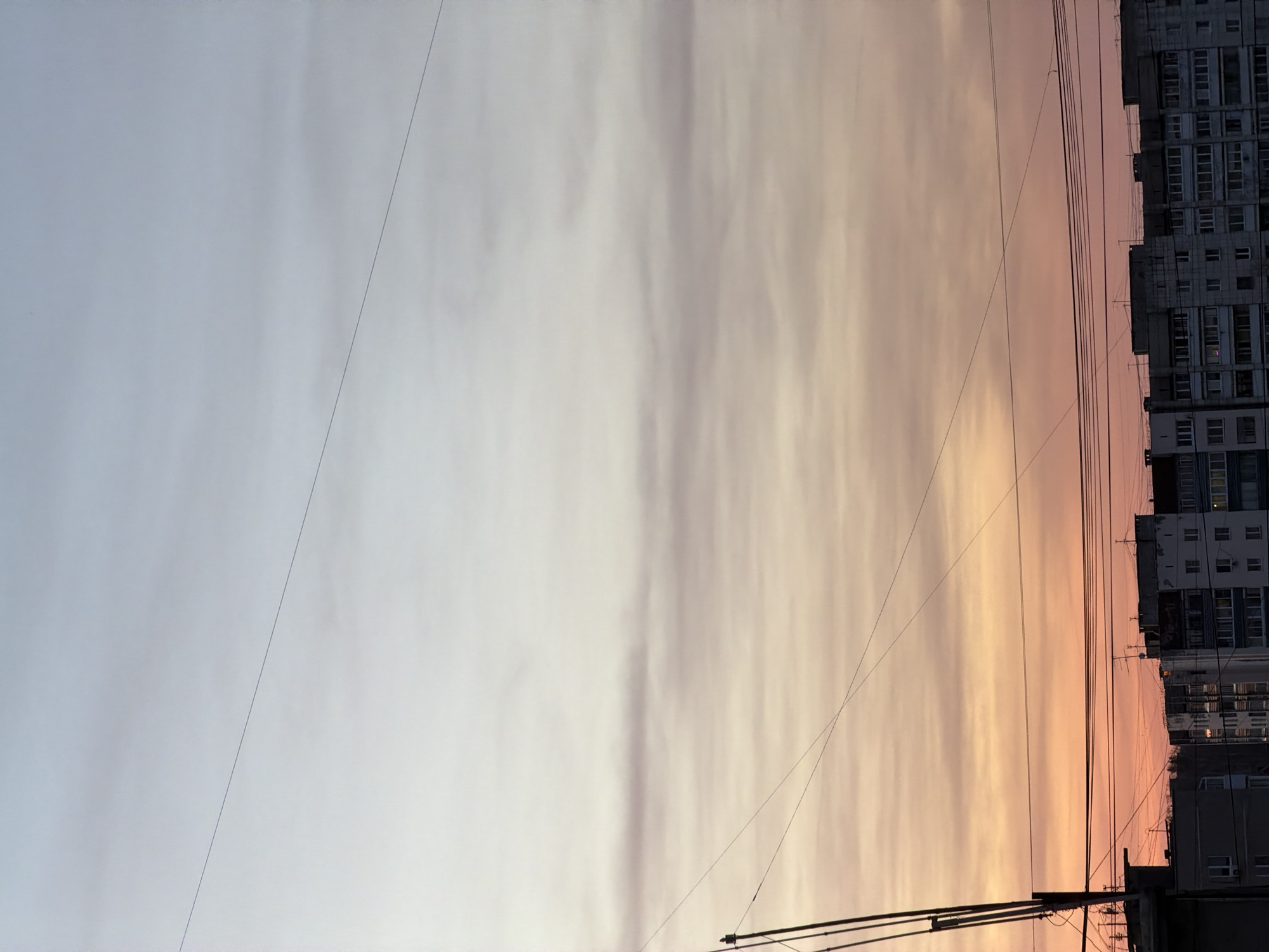 This vertical photograph captures an urban landscape during either sunrise or sunset in Perm, Russia. The expansive sky dominates the upper portion of the frame, showcasing a striking color gradient from a cool, light blue-gray on the left to a warm, vibrant orange and pink on the right. Delicate, streaky cloud formations are subtly visible across the sky, catching the soft, diffused light.
On the right side of the image, a row of dark, multi-story apartment buildings forms the urban horizon. These utilitarian structures, characteristic of post-Soviet architecture, feature numerous windows and appear somewhat weathered. Several thin, dark power lines or utility cables crisscross the sky, particularly in the lower right, creating geometric patterns against the natural backdrop and connecting to unseen utility poles at the very bottom edge of the frame.
The image portrays a serene moment in an urban setting, highlighting the atmospheric beauty of the sky contrasting with the man-made structures. The soft, directional light strongly suggests either the early morning or late evening hours. No people or visible text are present in the photograph.