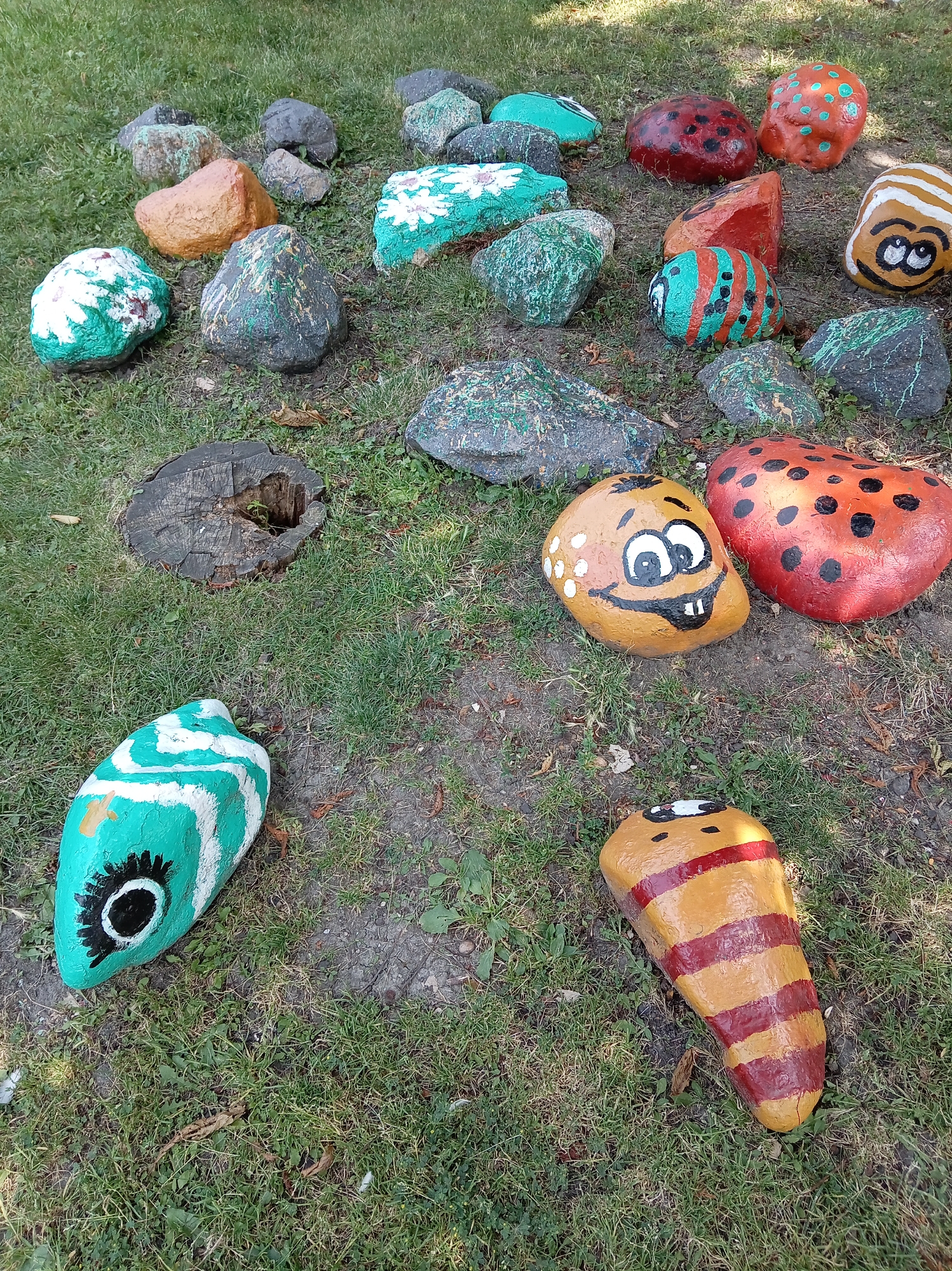 The image displays a collection of painted rocks scattered across a grassy area. Some rocks are painted to resemble ladybugs, with red and black dots. Others are decorated with various patterns and colors, including green with white flowers, and a fish-like shape with turquoise and white stripes and a large eye. One rock is painted to look like a cartoonish face with a wide grin and big eyes. Another rock has horizontal stripes in red and yellow. In the background, there are unpainted natural rocks and a tree stump. The scene is outdoors, with natural light illuminating the painted rocks. The environment suggests a park or garden setting. No people are visible in the image. The location is Yessentuki, Russia.