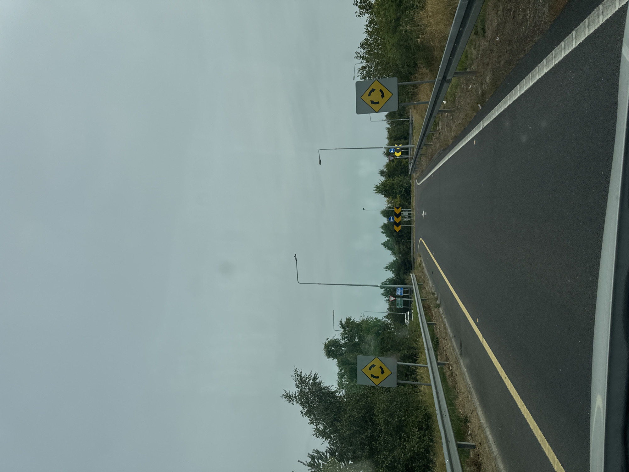 This is a first-person perspective, eye-level, outdoor, daytime, medium shot of a road in Father Campion Place, Ireland. The road is asphalt and has a yellow line on the left and a white line on the right. There are guardrails on both sides of the road. On the left side of the road, there is a yellow diamond-shaped sign with black arrows indicating a roundabout. Further down the road, there are more signs, including directional signs. On the right side of the road, there is another yellow diamond-shaped sign indicating a roundabout. The sky is overcast and appears light blue-gray. Trees and vegetation are visible on the sides of the road. The scene appears to be a highway or major road leading towards a roundabout.