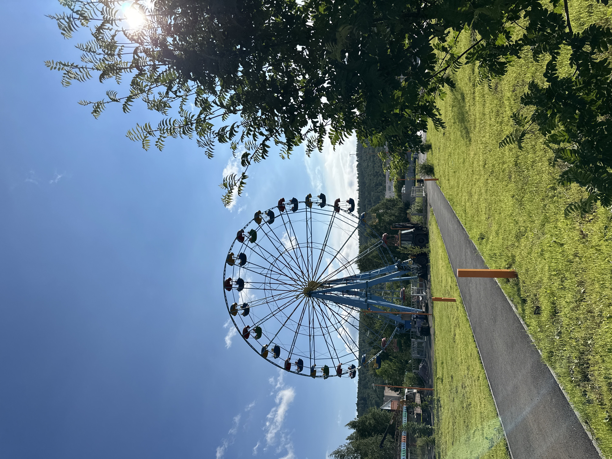 This is a low-angle, vertical shot of a Ferris wheel on a sunny day. The Ferris wheel, with its blue frame and colorful cabins, dominates the center of the frame. It appears to be in an amusement park or recreational area, as suggested by the grassy hills and trees in the background, as well as a paved path leading towards the Ferris wheel. The sun is bright, creating lens flare through the leaves of a tree in the upper left corner of the image. The sky is a clear blue with scattered white clouds. The lighting suggests it is daytime, likely late morning or early afternoon. There are no people visible on the Ferris wheel or in the immediate vicinity, giving the scene a sense of quiet anticipation. The overall mood is cheerful and summery, evoking a sense of fun and leisure. There is no visible text in the image.