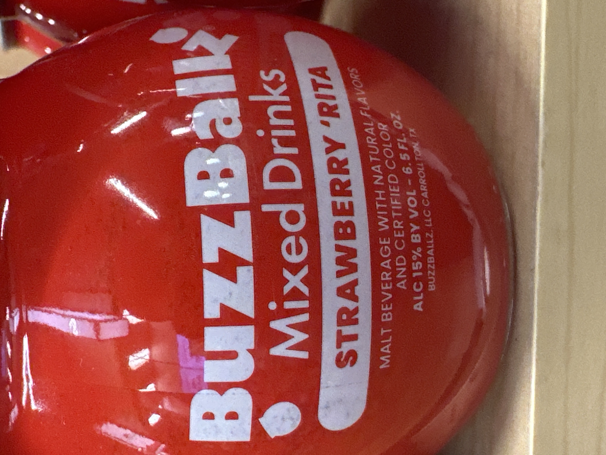 A red, round bottle of "BuzzBallz Mixed Drinks" is visible. The label on the bottle is white and reads "BuzzBallz Mixed Drinks Strawberry 'Rita" and includes nutritional information. The bottle is sitting on a wooden shelf. There are reflections of lights and other objects on the surface of the bottle. The image was taken in Camp Hill, United States of America.