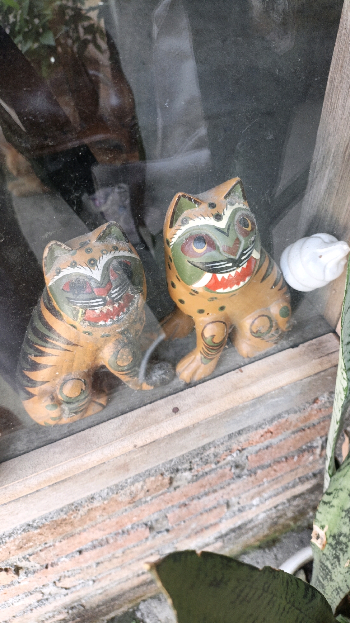 Two decorative figurines resembling tigers are positioned behind a glass pane. The figurine on the left is facing forward, while the one on the right is angled slightly to the right. Both figurines are painted in shades of orange and black, with green and red accents on their faces, depicting stylized tiger features such as stripes, open mouths with teeth, and wide eyes. The background, visible through the glass, is blurred and appears to be an indoor setting with foliage and reflections. To the right of the figurines, a small, white, shell-like object is visible. The foreground shows a wooden ledge with brickwork below it, and a glimpse of a green plant leaf. The overall scene suggests these figurines are displayed on a shelf or windowsill. The location in Cikal, Indonesia, is not discernible from the visual information in the image.