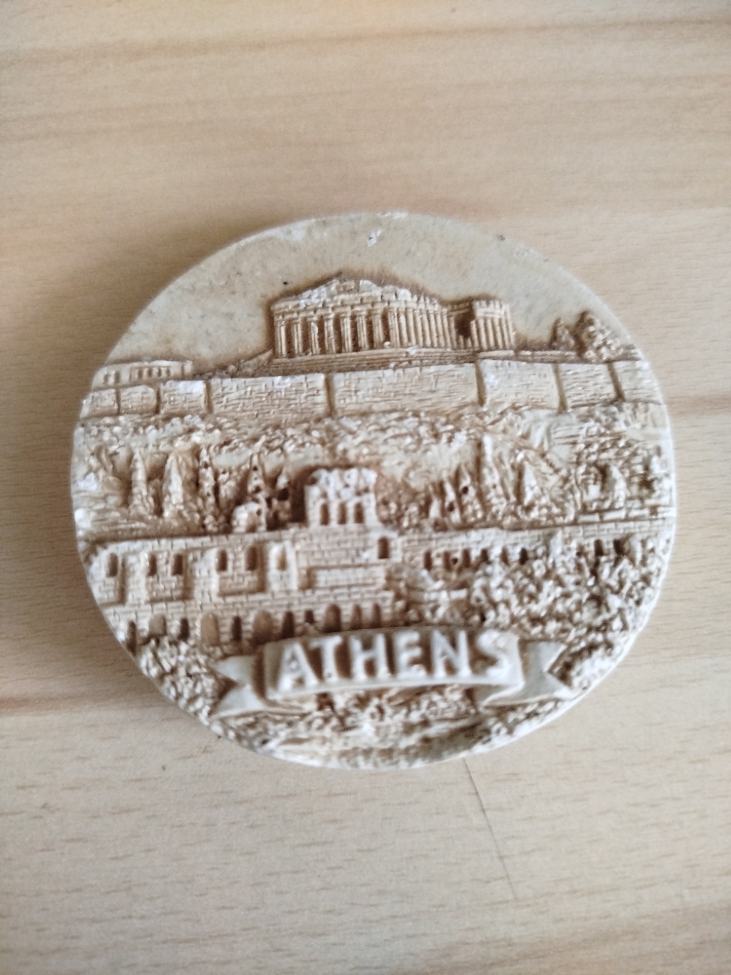 The image shows a circular, beige-colored souvenir depicting a scene from Athens, Greece. The Parthenon is visible at the top, above a fortification wall. Below the wall are other ancient buildings and foliage. A banner at the bottom reads "ATHENS". The souvenir is lying on a light brown, wood-grained surface. There is no evidence that this photo was taken in Varna, Bulgaria.