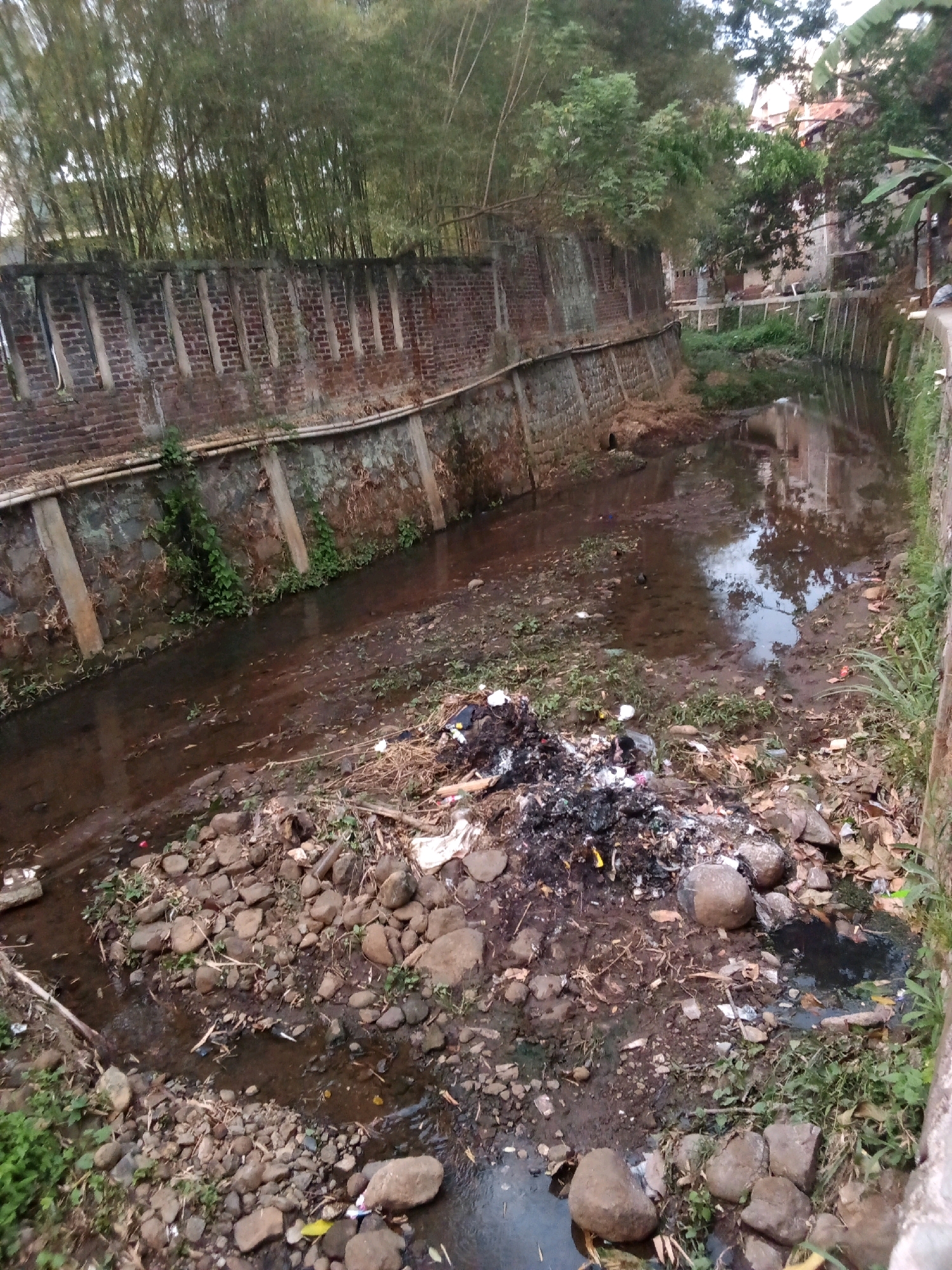 The image depicts a narrow, dirty canal in an urban environment, likely in Bandung City, Indonesia. The canal walls are constructed of brick and concrete, with some vegetation growing from the cracks. The water in the canal is brown and stagnant, with a significant amount of debris floating on the surface. Piles of garbage, including plastic bags, paper, and other refuse, are visible on the banks of the canal and in the water. Bamboo trees and other greenery line the top of the canal walls, suggesting a location with some natural elements within the city. The overall scene conveys a sense of neglect and pollution.
