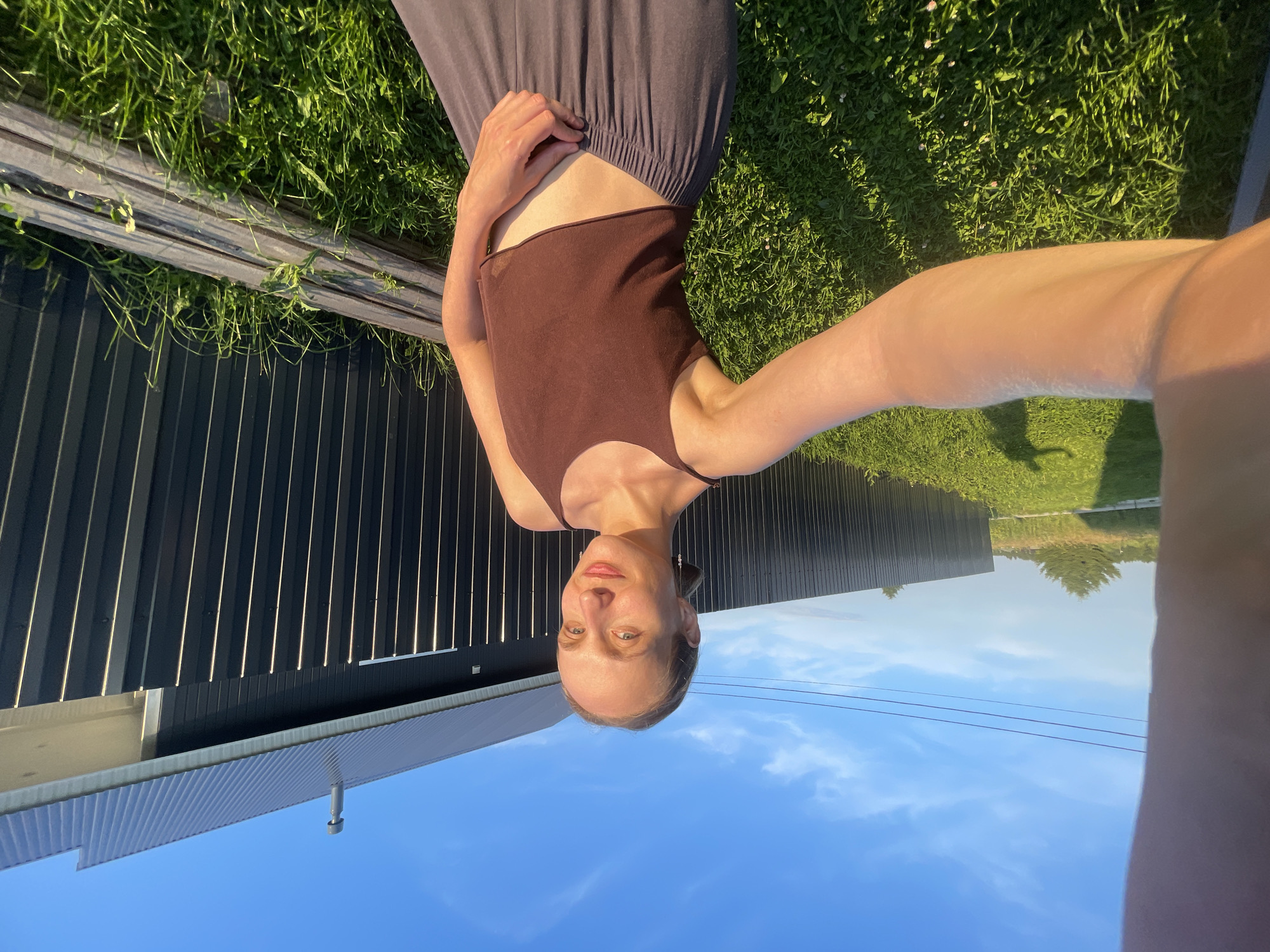 The media file shows a young woman taking a selfie outdoors. She is wearing a brown tank top and a dark grey skirt. The woman is standing on grass in what appears to be a backyard, with a dark corrugated fence behind her and a clear blue sky above. The lighting suggests it is late afternoon or early evening, with the sun casting long shadows. The overall mood is relaxed and casual. The image is oriented sideways. There is no visible text. The location is Podshivalovo, Russia.