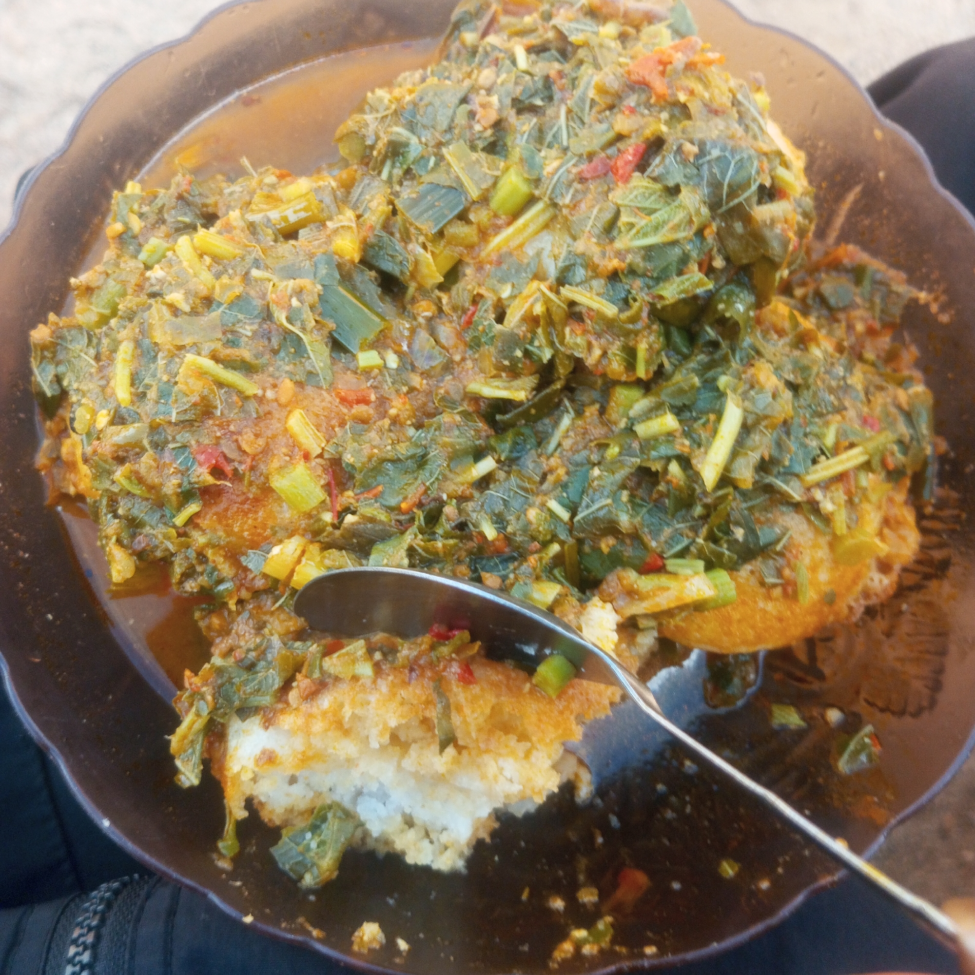 A close-up, overhead shot shows a plate of food, likely fufu and a leafy green stew, served in a dark brown, scalloped plastic bowl. A silver spoon is partially submerged in the food, lifting a portion of the stew to reveal white fufu underneath. The stew is a mix of chopped green leaves, some red pepper flakes, and yellow pieces, all coated in a reddish-brown sauce. The bowl rests on a dark surface, possibly fabric. The background is out of focus, suggesting an outdoor setting in Jos, Nigeria, given the commonality of this dish and the environmental cues. The lighting is natural, indicating daytime.