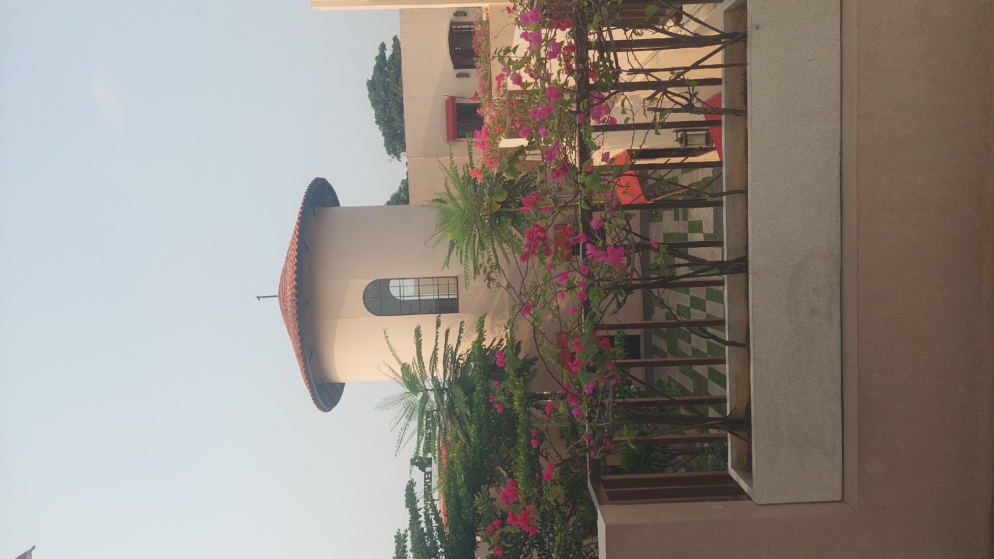 A close-up shot captures a building with a cylindrical tower, a tiled roof, and a large arched window. In the foreground, a planter box holds several small trees. Behind the planter, a dark metal fence encloses a checkered patio area. Bright pink bougainvillea flowers are abundant throughout the scene. The building is painted a pale beige color, with red awnings visible over some windows. The sky is overcast and light blue. The scene is located in Cileungsi, Indonesia.