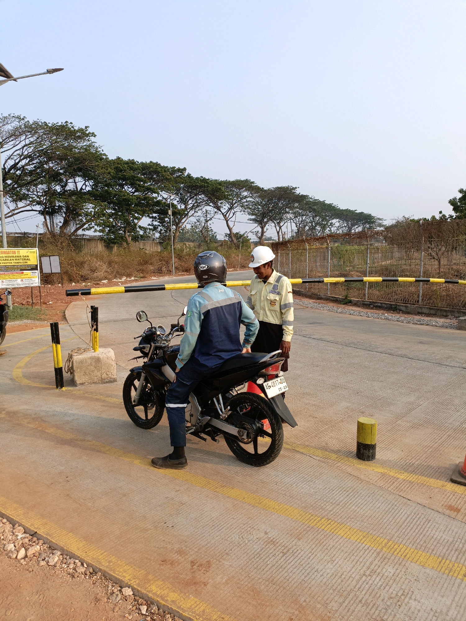 The image depicts an outdoor scene at what appears to be a checkpoint or entrance in Jenu, Indonesia. In the foreground, a person wearing a blue and grey uniform, a dark helmet, and dark pants is seated on a black motorcycle. The motorcycle's license plate reads "AE 4017 DU". To the right of the motorcycle, another person wearing a light yellow uniform with reflective stripes, dark pants, and a white hard hat stands and appears to be interacting with the person on the motorcycle. A yellow and black striped barrier gate is lowered across the road behind the two individuals. The environment includes a paved road marked with yellow lines, gravel, some vegetation, trees in the background, and a fence. A lamppost is visible in the upper left corner. A sign with Indonesian text is mounted on a post to the left of the barrier. The sky is clear and appears to be late afternoon.