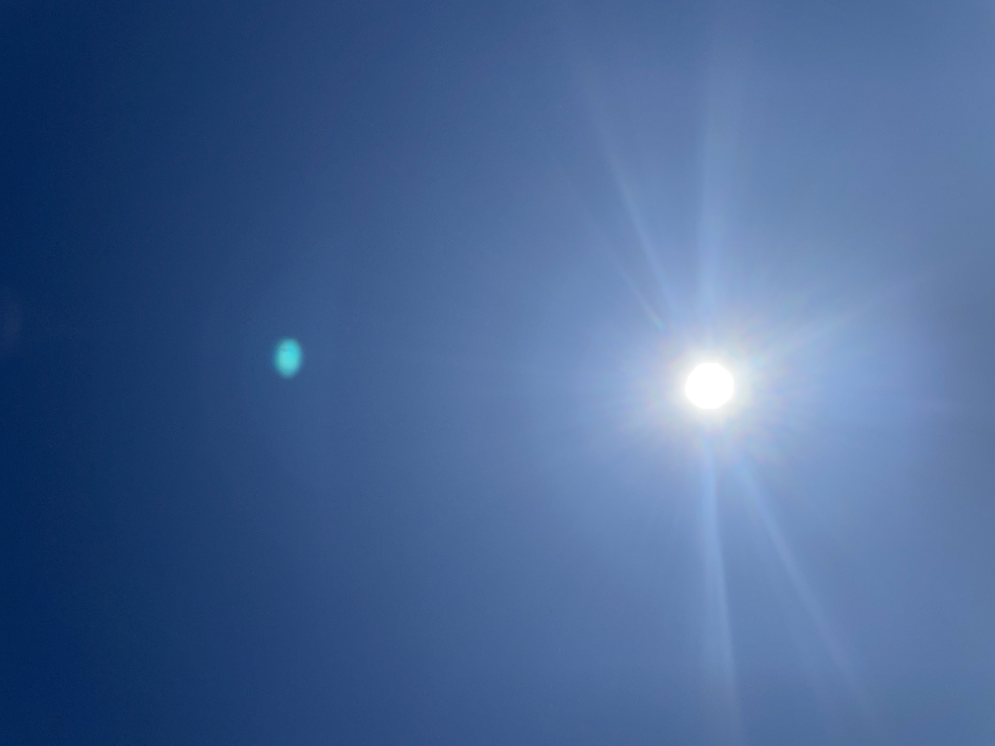 The image depicts a bright, clear blue sky with the sun visible in the upper right portion of the frame. A small, out-of-focus, light blue/green circular artifact is visible in the left portion of the frame. The scene appears to be a simple view of the sky, without any identifiable features to connect it to Поедуги, Russia or any other specific location. No people, subjects, objects, actions or interactions are present in the scene, beyond the celestial and optical phenomena.