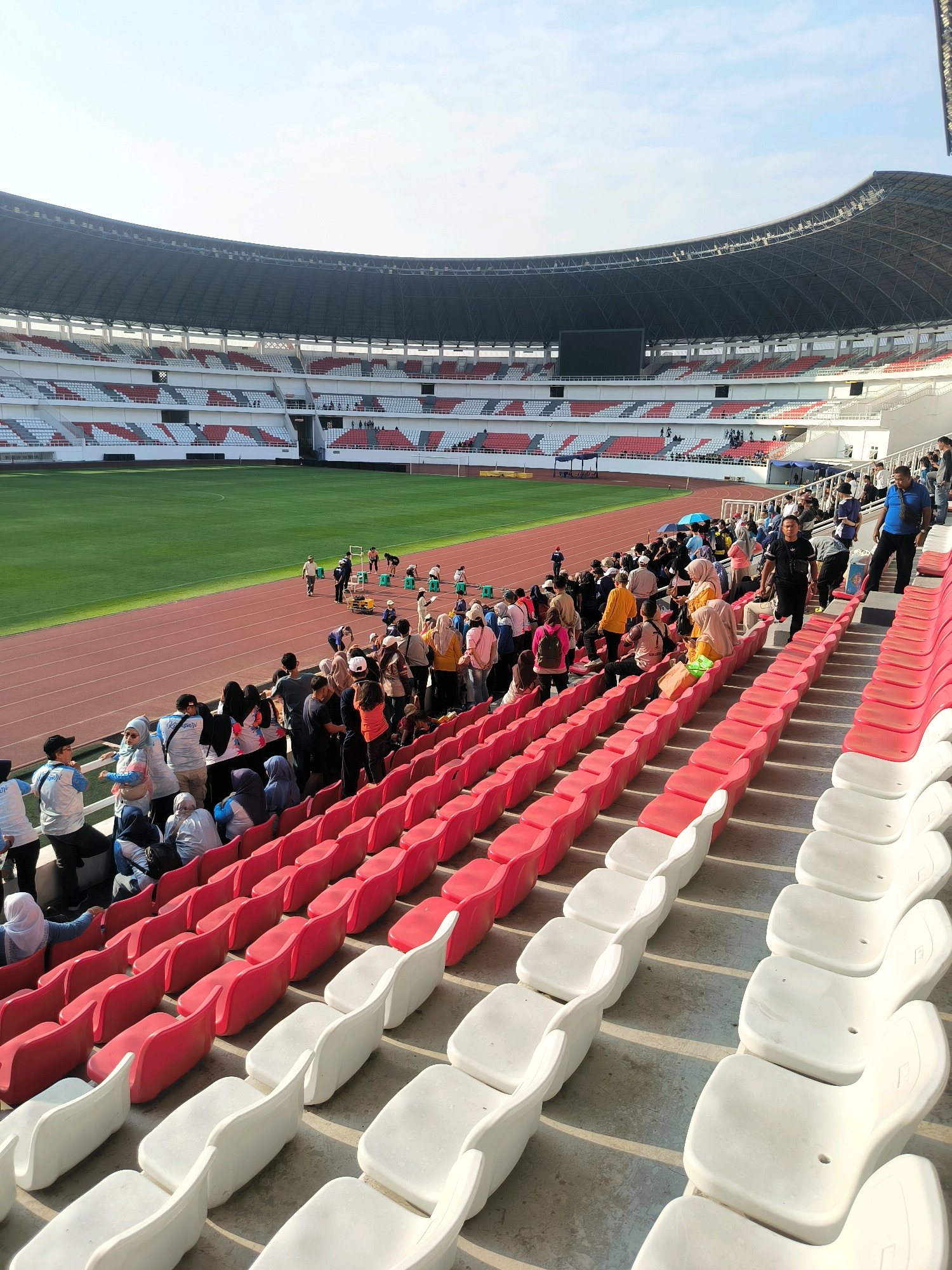 The image shows a bright, daytime view of the Gelora Jatidiri Stadium in Semarang, Indonesia, a multi-purpose stadium with a large capacity. The stadium features a green grass field with running tracks surrounding it, a vast seating area with alternating red and white seats, and a covered grandstand. A significant number of people, likely visitors or participants, are gathered in the stadium, some standing along the railings overlooking the field, while others are seated or milling about in the stands. The group appears diverse, with many individuals wearing casual clothing and some wearing headscarves, suggesting a varied crowd. The atmosphere seems relaxed and organized, with people moving about and observing the surroundings. The stadium's architecture is modern, with a large roof structure visible in the background. The sunlight casts shadows across the seating area, indicating a clear day.