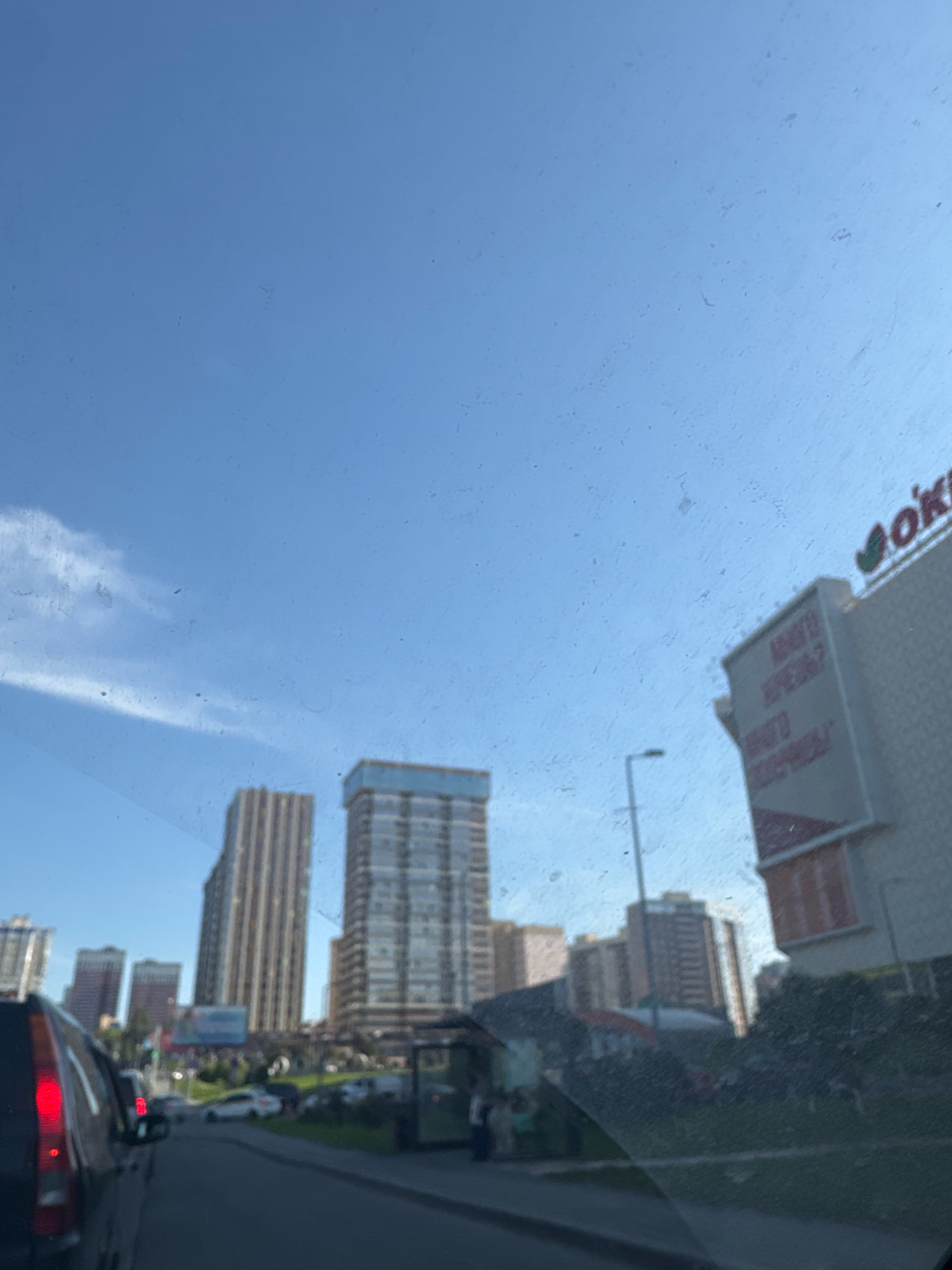 The image depicts a street scene in Novosibirsk, Russia, on a clear day with a bright blue sky. In the foreground, the left side of the frame shows the rear of a dark-colored SUV, its brake lights illuminated. The road ahead is lined with buildings, including several tall apartment buildings and a larger commercial building with signage visible. One sign on the commercial building appears to read "Oki." Further down the street, a bus stop shelter is present, and several cars are parked or driving on the road. The scene suggests a typical urban environment, possibly during daylight hours.