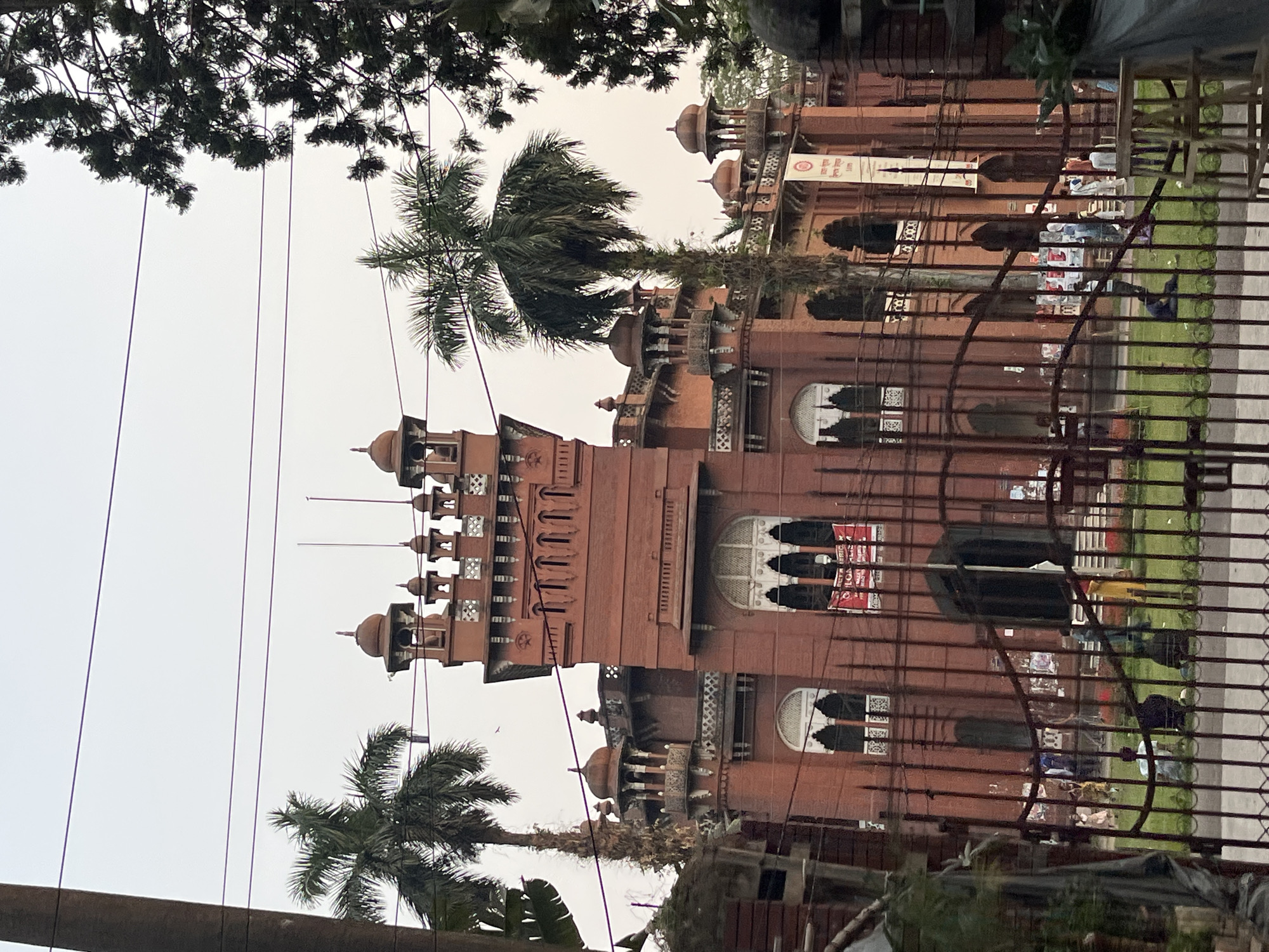 The image shows Curzon Hall at the University of Dhaka in Dhaka, Bangladesh. It is a large, red brick building with towers, domes, and arched windows, partially obscured by a fence and trees. People are visible on the lawn in front of the building. Overhead electrical wires intersect the view of the building.