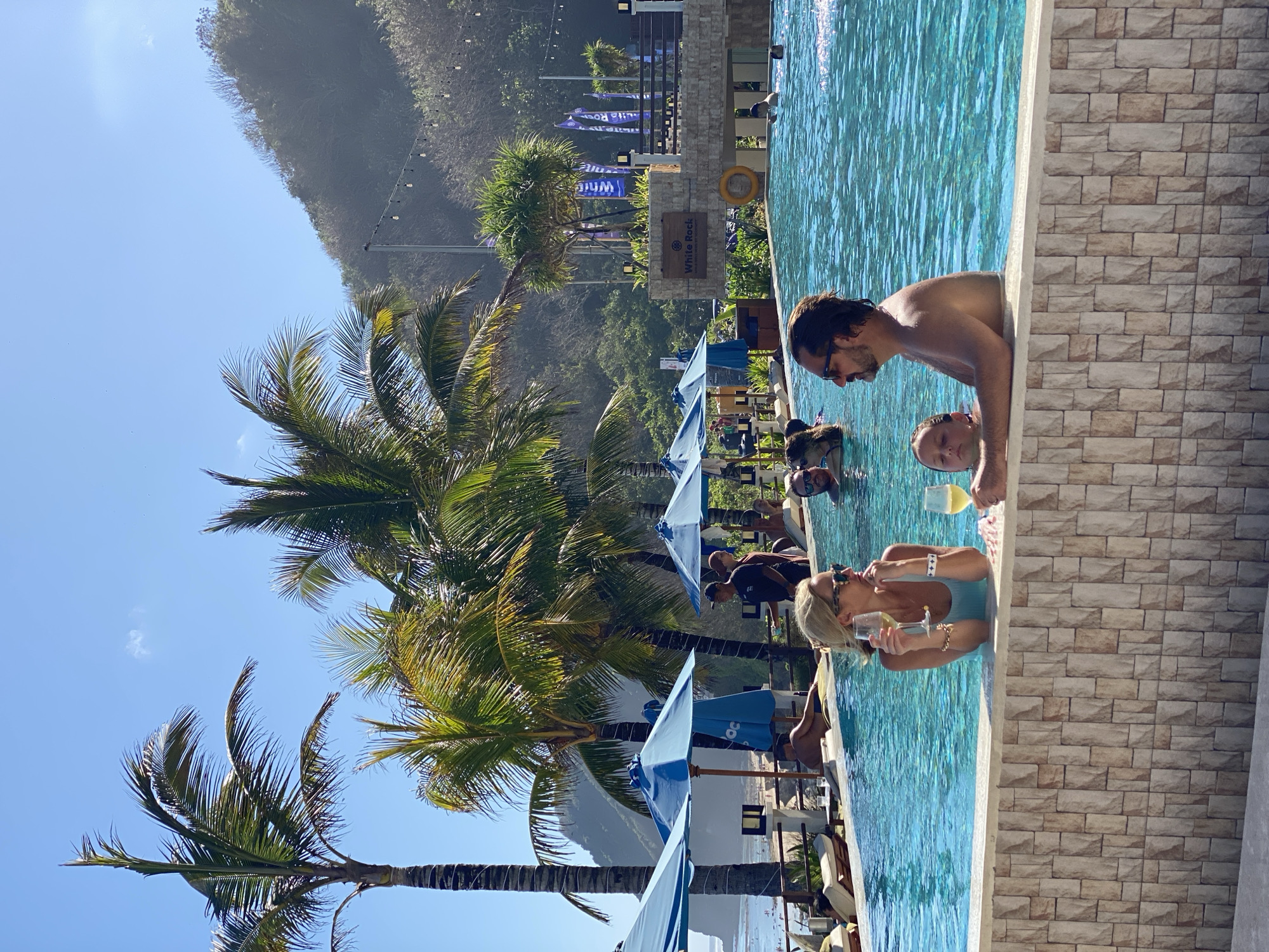 In Kuta Selatan, Indonesia, a resort pool is occupied by several people. In the foreground, a couple stands in the pool, each holding a glass of white wine. The woman, with blonde hair and sunglasses, wears a light blue swimsuit. The man, with dark hair and sunglasses, faces left. To their right and slightly behind them, a young girl with blonde hair is also in the pool. In the middle ground, other individuals are visible in the pool, with palm trees, blue umbrellas, lounge chairs, and a distant beach providing a tropical backdrop. A building with signage reading "White Rock" is situated behind the pool area, further enhancing the resort atmosphere. The scene is bathed in bright sunlight, suggesting a clear, warm day.