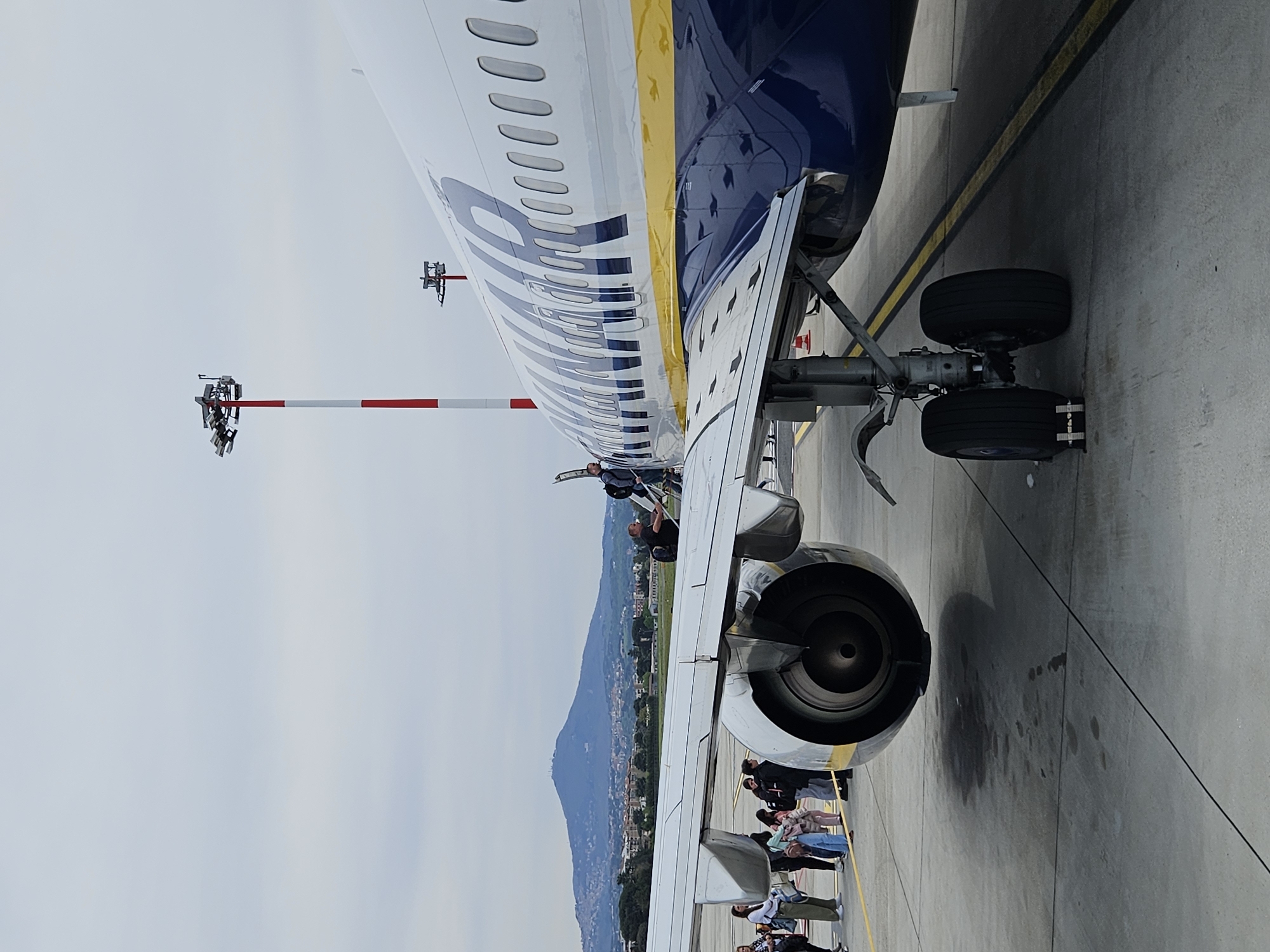 The image shows a Ryanair airplane on the ground, with passengers boarding via a set of stairs attached to the plane's wing. The plane is parked on an asphalt apron, and the ground is marked with black lines. A small group of people is visible boarding the plane. In the background, there is a view of a mountain and a cityscape that is similar to Rome, Italy. A red and white striped pole with lights or sensors is visible near the front of the plane.