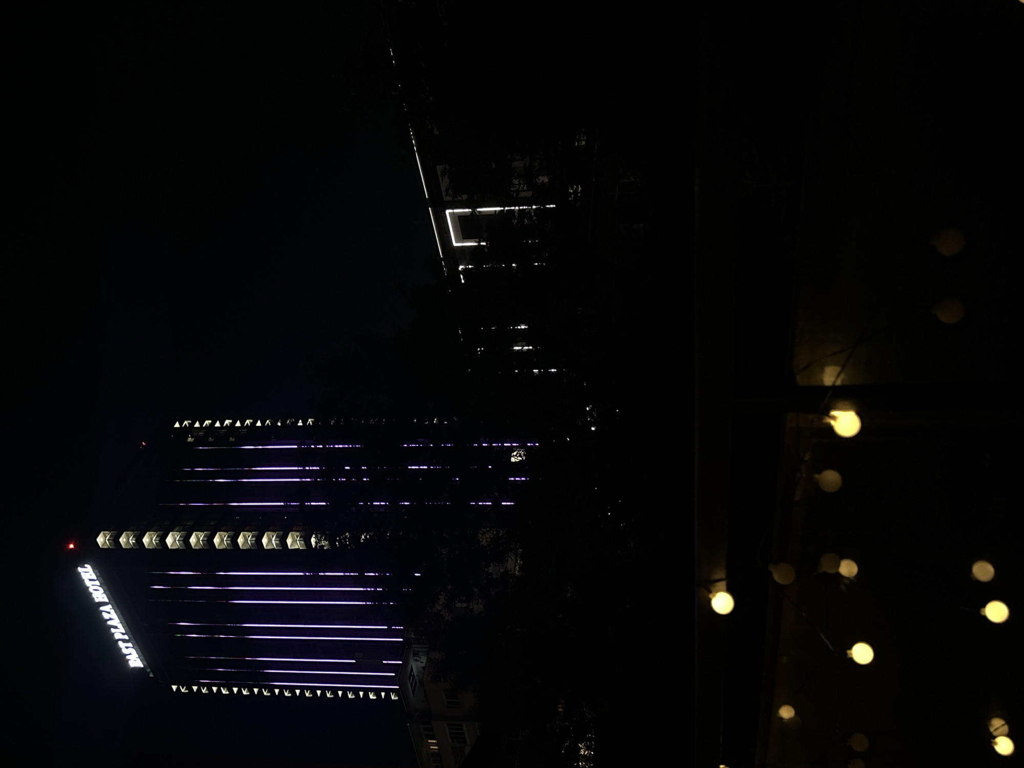 The image shows a nighttime view of the East Plaza Hotel in Sofia, Bulgaria, with its facade illuminated by purple vertical lights. The hotel is a tall, modern building with numerous floors and balconies. To the right of the hotel, another building is visible, also illuminated by lights. In the foreground, a string of yellow lights is blurred, suggesting they are close to the camera and out of focus. The overall scene is dark, with the exception of the illuminated buildings and the foreground lights.