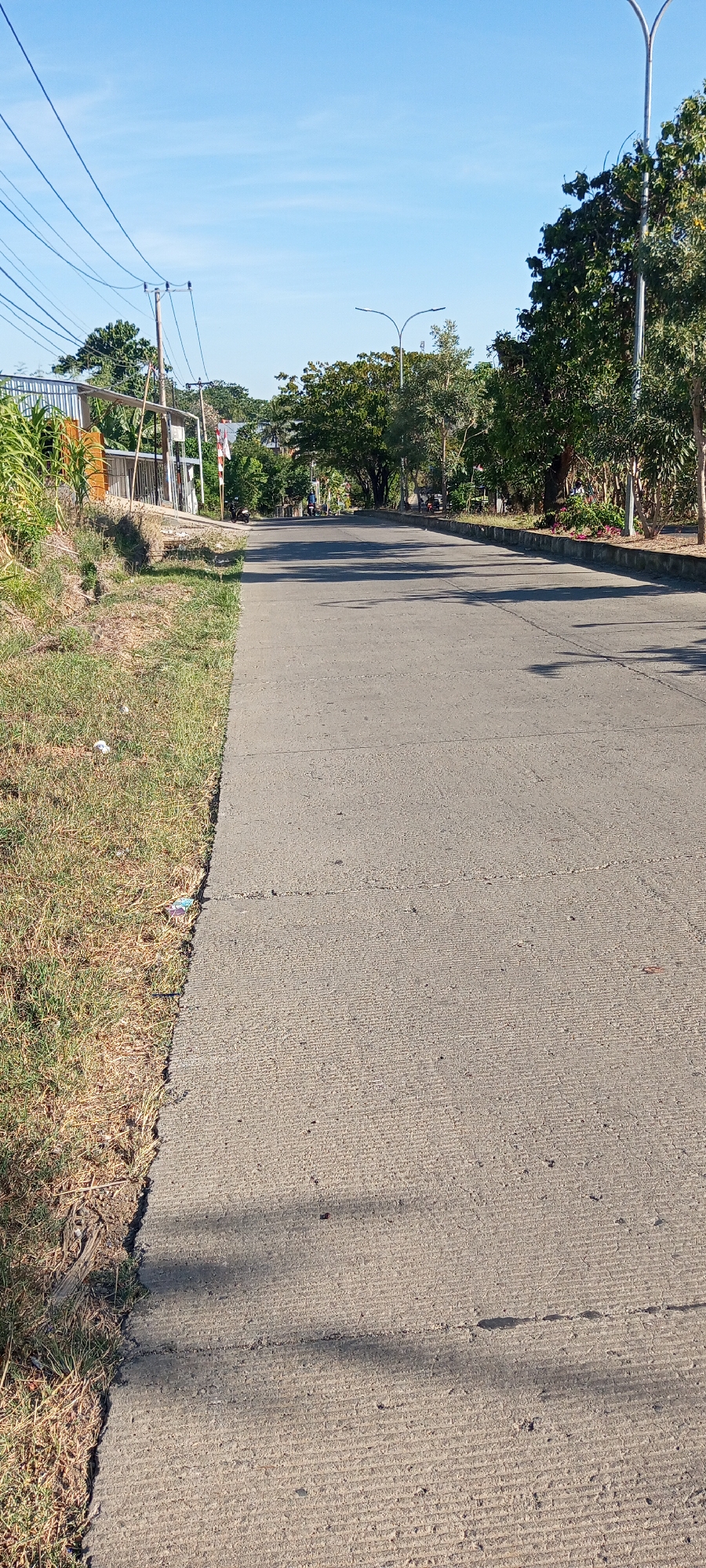 The image shows a paved road in Bulupabbulu, Indonesia, under a clear blue sky. The road is bordered by a grassy verge on the left and trees on the right. Power lines stretch across the sky, supported by utility poles. In the distance, several motorcycles and people are visible, suggesting moderate activity. Buildings and more trees can be seen further down the road. The scene appears to be a typical street view in a residential or semi-rural area.
