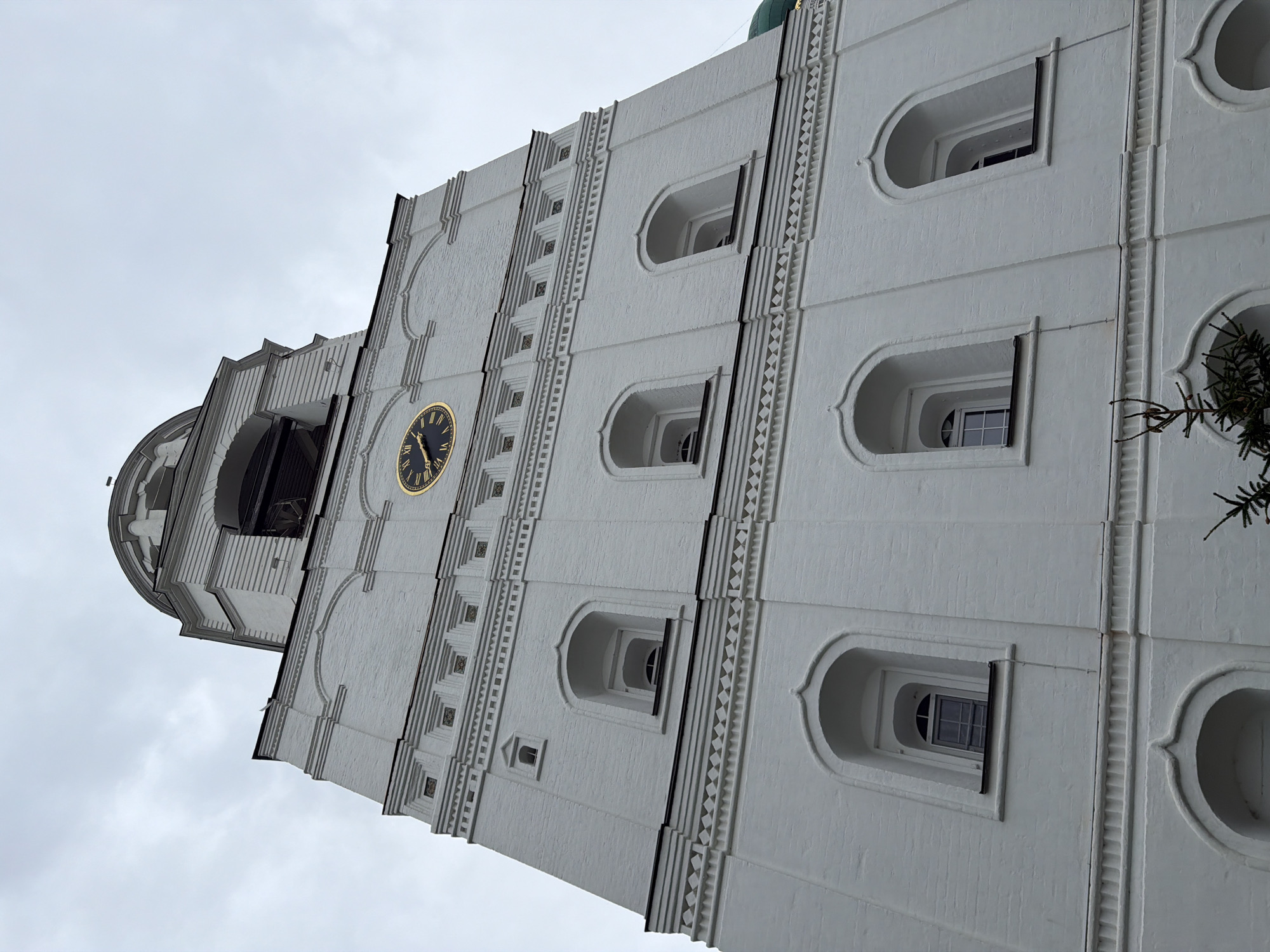 This is a low-angle, close-up, full shot of the white brick bell tower of the Church of the Assumption of the Blessed Virgin Mary in Rusanovo, Russia. The tower is tall and rectangular with decorative architectural elements. It features multiple tiers of arched windows. A large, round clock with Roman numerals and gold hands is visible near the top of the tower. Above the clock, there are openings where bells would be located, and a domed roof with a cross at the very top. The sky is overcast and grey. A small evergreen branch is visible in the lower right corner.