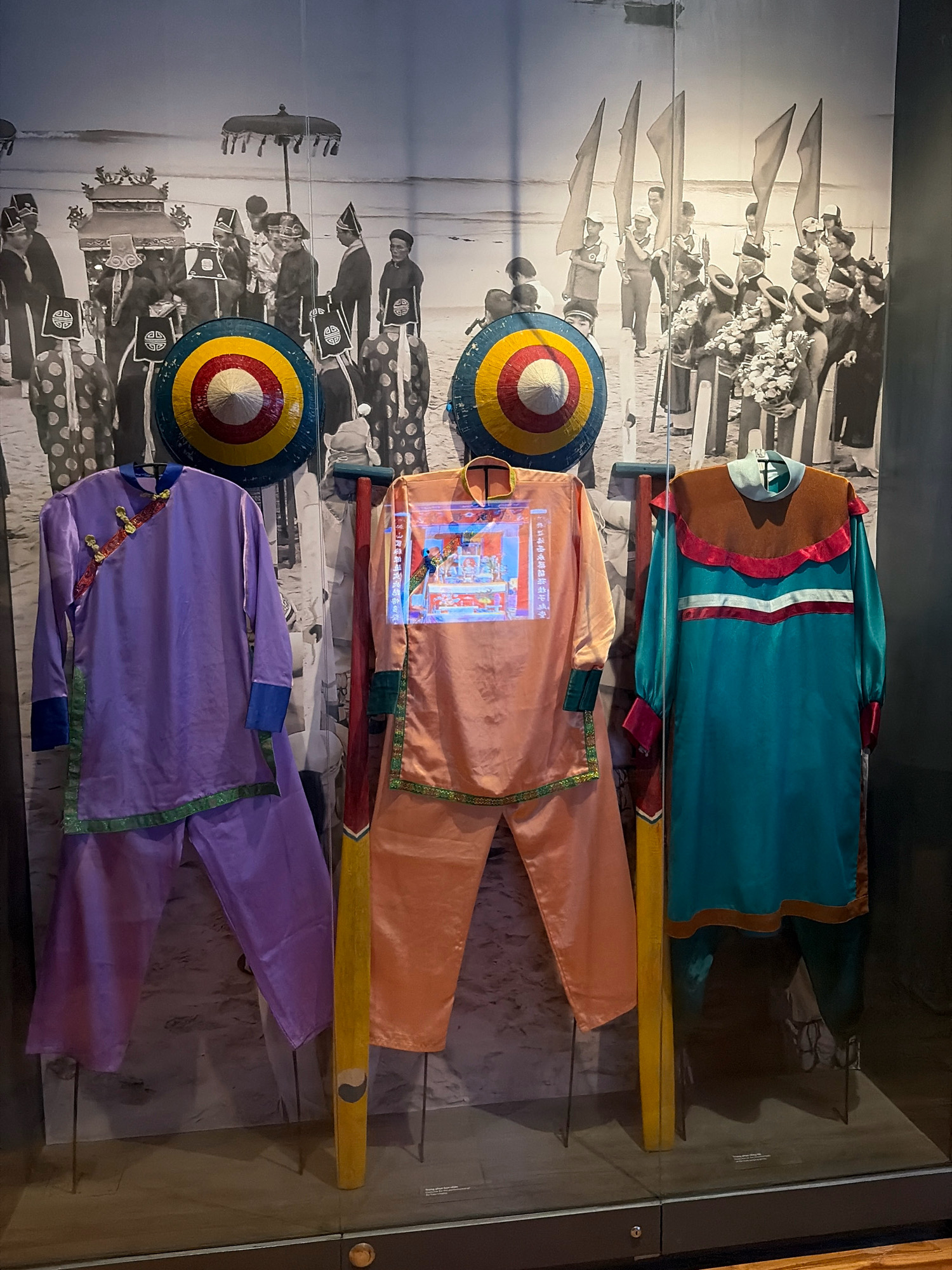 The image shows a display in a museum in Da Nang, Vietnam. The display features three traditional Vietnamese outfits on mannequins. The outfit on the left is purple with green trim and blue cuffs. The outfit in the middle is peach-colored with green trim and a colorful projected image of what appears to be a religious ceremony on its chest. The outfit on the right is teal with brown and red trim. Behind the outfits is a black and white mural depicting a procession or ceremony with many people in traditional attire. Two conical hats with colorful targets are also visible, positioned in front of the mural. The outfits are displayed on thin metal stands. The flooring is a neutral color, and the display case is made of glass.