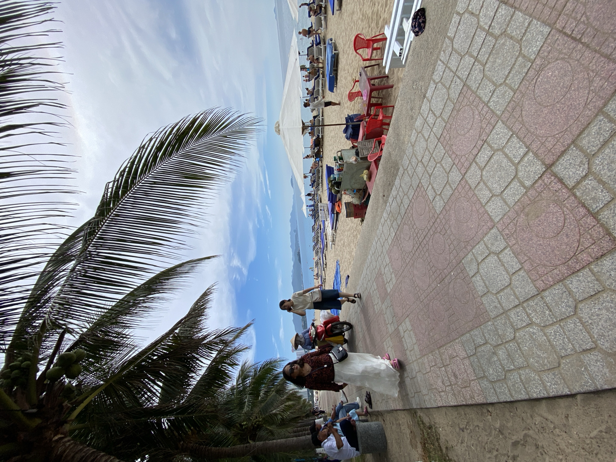 A beachfront scene in Nha Trang, Vietnam, features a paved walkway in the foreground with a patterned surface of pink and gray square tiles, bordered by sand on one side. Several palm trees with long fronds arch over the scene from the left. On the walkway, two women are walking: one in the foreground wearing a dark patterned top, a long white skirt, and sunglasses, and another woman with a white top, dark shorts, and a crossbody bag. Behind them, a person wearing a conical hat rides a red scooter, accompanied by another individual. Further down the walkway, several people are seated, some under the shade of palm trees. Beyond the walkway lies a sandy beach populated with numerous beach chairs and umbrellas, some occupied by beachgoers. In the distance, the calm blue sea stretches towards a hazy horizon with a mountainous coastline. The sky above is a mixture of blue with scattered white clouds. A small vendor cart is visible on the sand, with coconuts and other items displayed.