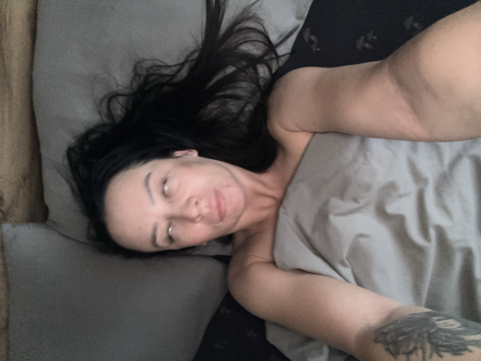 A woman lies in a bed. Her head rests on a gray pillow, and she is partially covered by a gray blanket. She has long dark hair spread out on the pillow. A tattoo is visible on her left forearm. The background includes dark clothing and a second pillow. There is no evidence to suggest that this image was taken in Chaikovsky, Russia.
