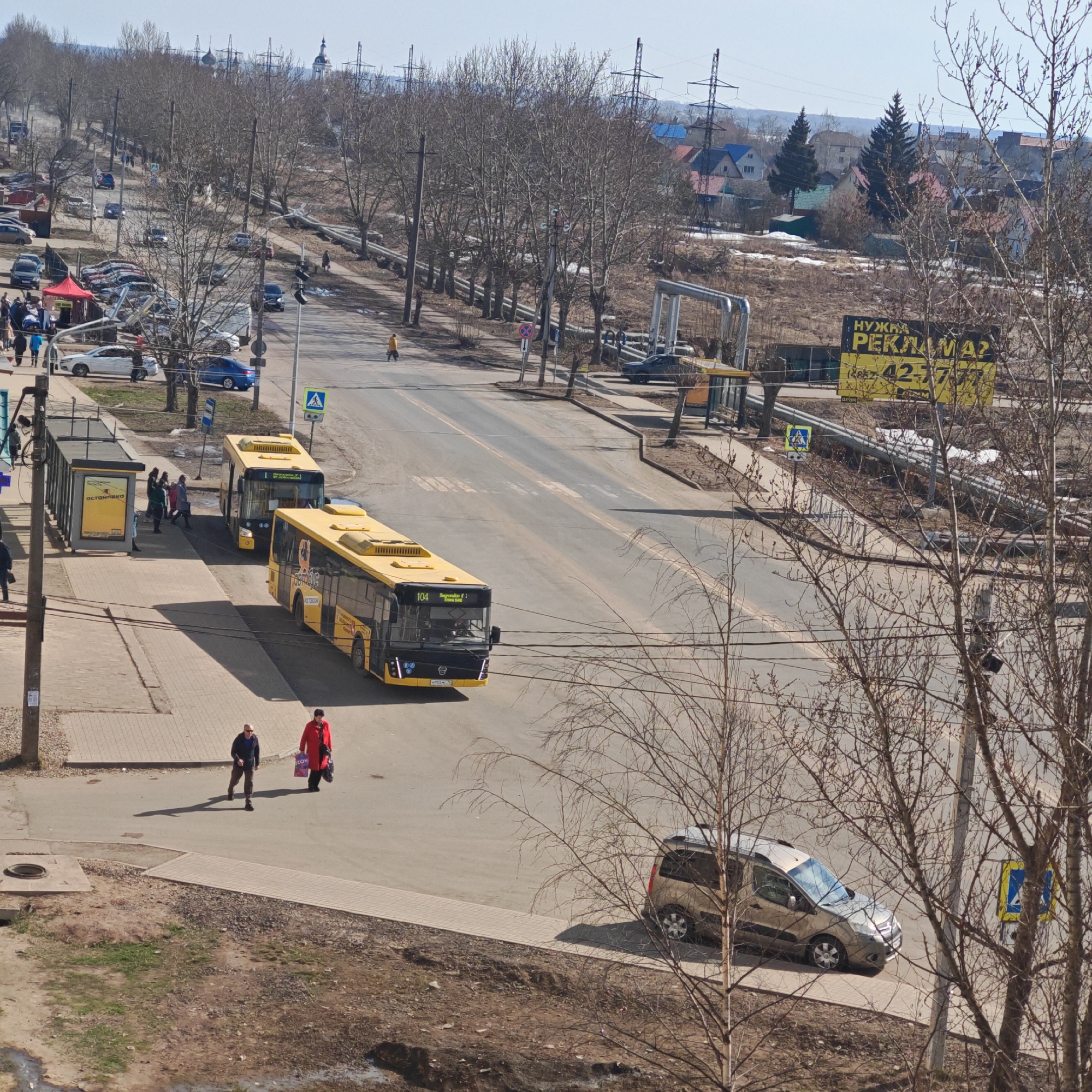 The image shows a street scene in what appears to be the city of Rostov, Russia. Two yellow buses are visible on the road, one closer to the camera with the number "104" displayed on its front. People are present at a bus stop. Two pedestrians, one wearing a red jacket, are walking on the sidewalk. A parked car is visible near a pedestrian crossing sign. Overhead power lines and bare trees line the street. In the background, buildings and a billboard with Russian text are seen.