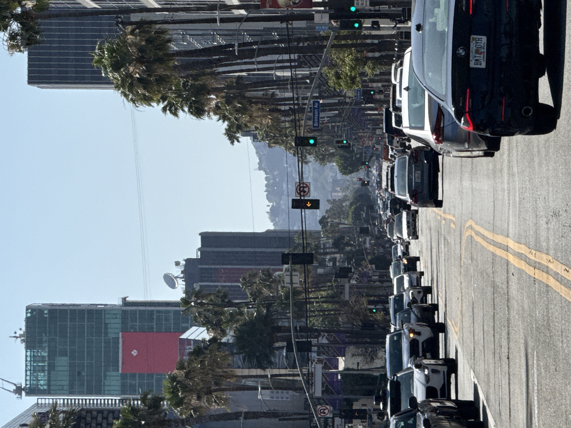 The image shows a congested street in Los Angeles, United States of America, lined with palm trees and tall buildings. A line of cars is stopped in traffic, facing forward. The street has double yellow lines, and signs for "Bronson Av" are visible. A building with a large red square on its side is visible in the background. A satellite dish is mounted on top of one of the buildings.