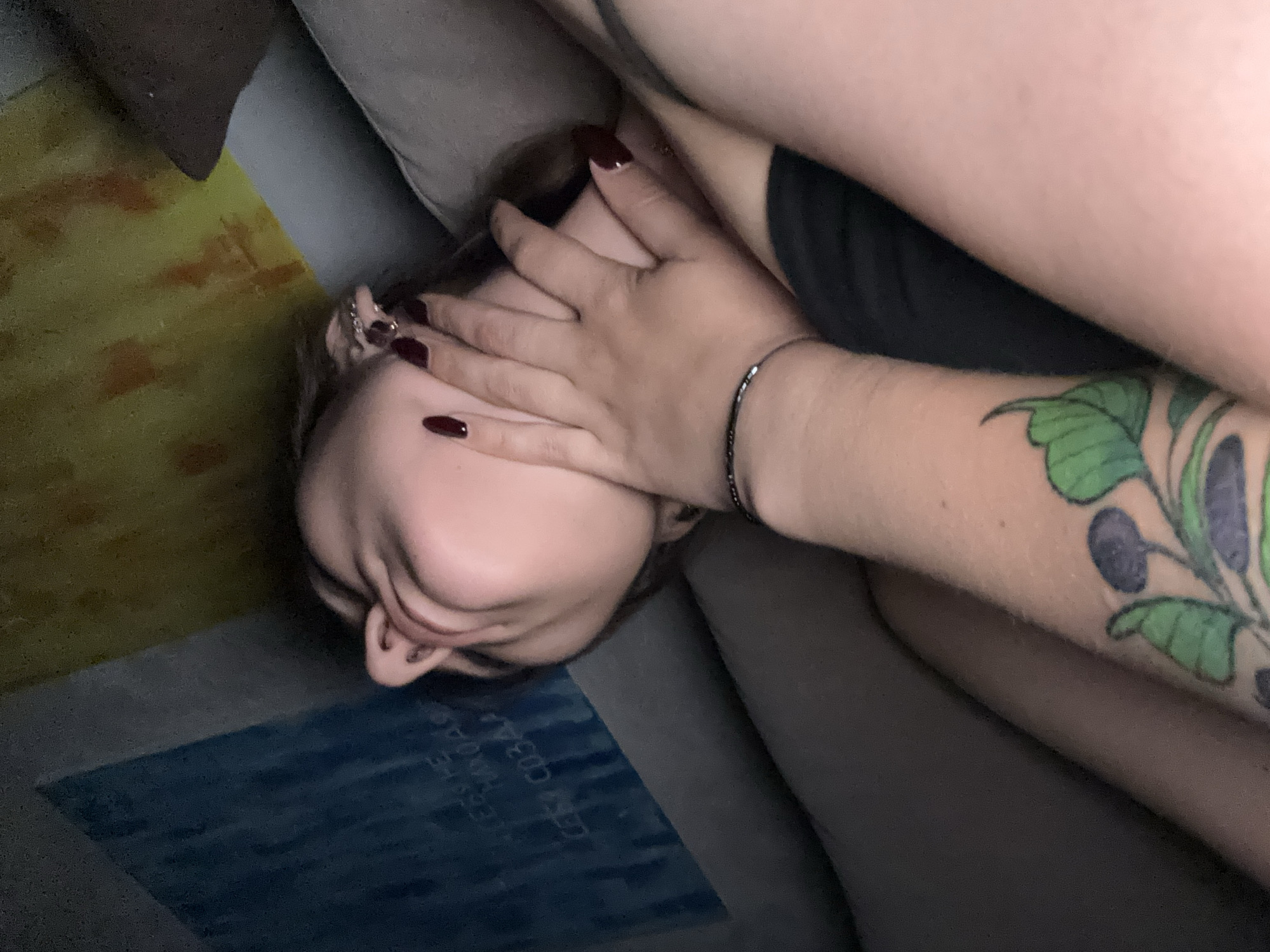 The image shows a close-up view of two people in what appears to be a domestic setting. One person, whose face is mostly obscured by a hand, is lying down. The hand covering their face has dark red nail polish and a thin black bracelet. The arm of the person whose hand is visible has a tattoo of green leaves and dark purple berries. The background includes a textured wall or fabric in shades of yellow and orange, and a blue surface with some faint text. The overall impression is intimate and casual. There is no information in the image to suggest the location is Novokuznetsk, Russia.