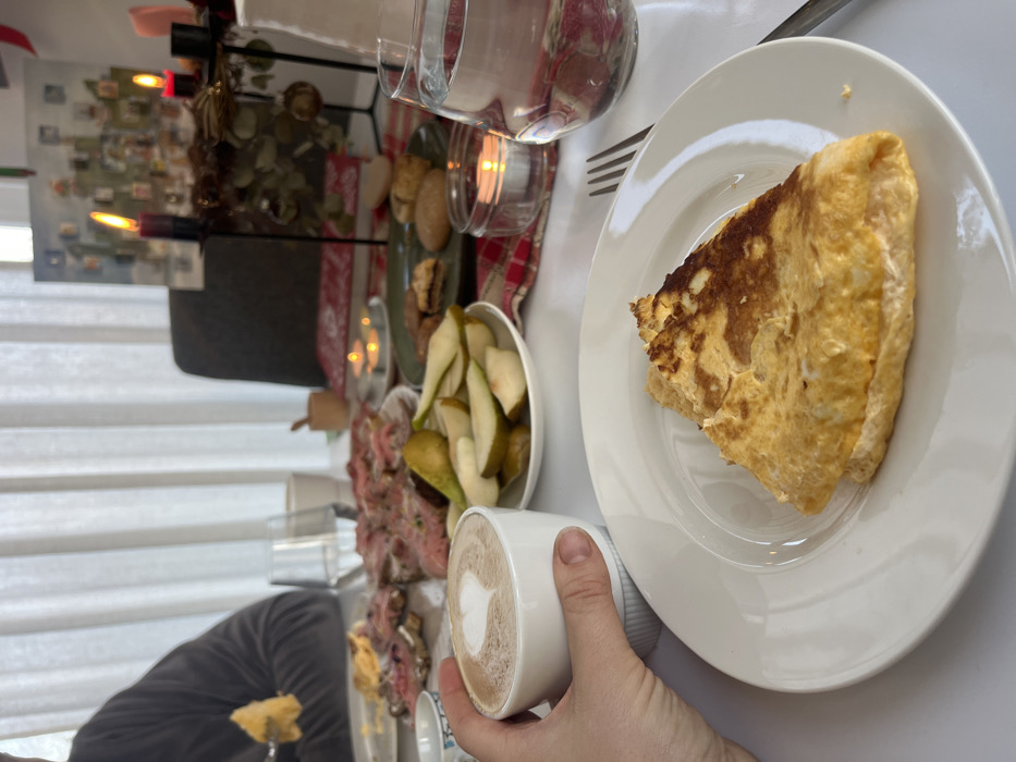 This image captures an indoor dining scene in Obermoschel, Germany, likely during a breakfast or brunch. The perspective is first-person, with a hand holding a white cup of coffee or latte featuring a heart-shaped latte art in the lower-left foreground. On a white plate directly in front of the viewer, a triangular portion of a cooked egg dish, resembling an omelette, is presented. A silver fork rests to the right of the plate.
The white table surface is laden with various food items and decorations. To the left of the viewer's plate, a white dish holds slices of green pears. Further back, a platter displays several open-faced sandwiches topped with thinly sliced pink meat, consistent with roast beef. A green plate to the right holds an assortment of cookies or biscuits. Two clear glasses filled with water are visible, one near the foreground plate and another further back. Decorative elements include a small glass votive with a lit tealight candle, a red and white plaid table runner, a small wooden mortar and pestle, and a red book.
In the mid-to-background, another individual is seated on the left, wearing a dark grey long-sleeved garment, actively eating a piece of the egg dish with a fork. Behind the table, an Advent candle holder with two lit black candles, adorned with natural elements and small ornaments, is prominently displayed. Further back, a large Advent calendar featuring numerous small doors or compartments is visible on what appears to be a wall. The background is dominated by white sheer curtains, suggesting a window and providing diffused natural light to the scene. A dark chair back is partially visible behind the table.