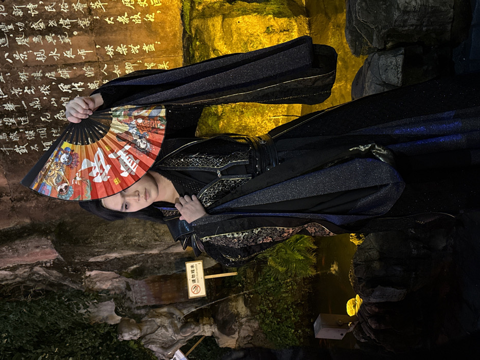 A person in a black, shimmering robe with ornate gold trim holds a colorful fan. The fan features illustrations and Chinese characters. The person stands near a rock formation, likely a historical site, given the inscribed Chinese characters on the rock face. A sign reading "请勿戏水 DON'T PUY MATER" is visible, suggesting the location may be near water. The setting appears to be a combination of natural rock formations with man-made decorative elements and lighting.
