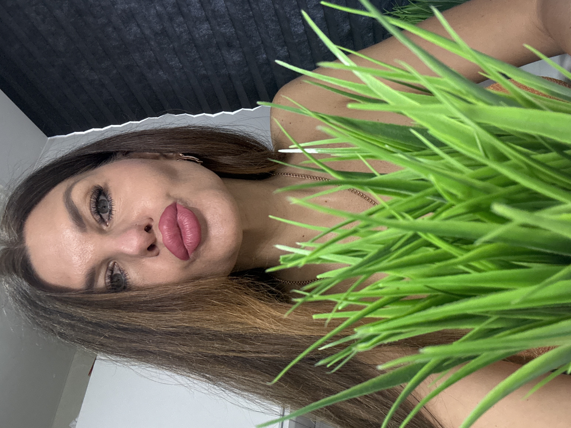 A medium shot, slightly rotated, shows a woman with long brown hair, light skin, and defined facial features including full lips, looking directly at the camera with a neutral expression. She appears to be indoors, as evidenced by a black, vertically ribbed backdrop in the upper left corner. Green, grassy plants are in the foreground, partially obscuring her body. A gold chain necklace is visible around her neck. The location is specified as Chaikovsky, Russia, though there are no visual cues in the image to confirm this.