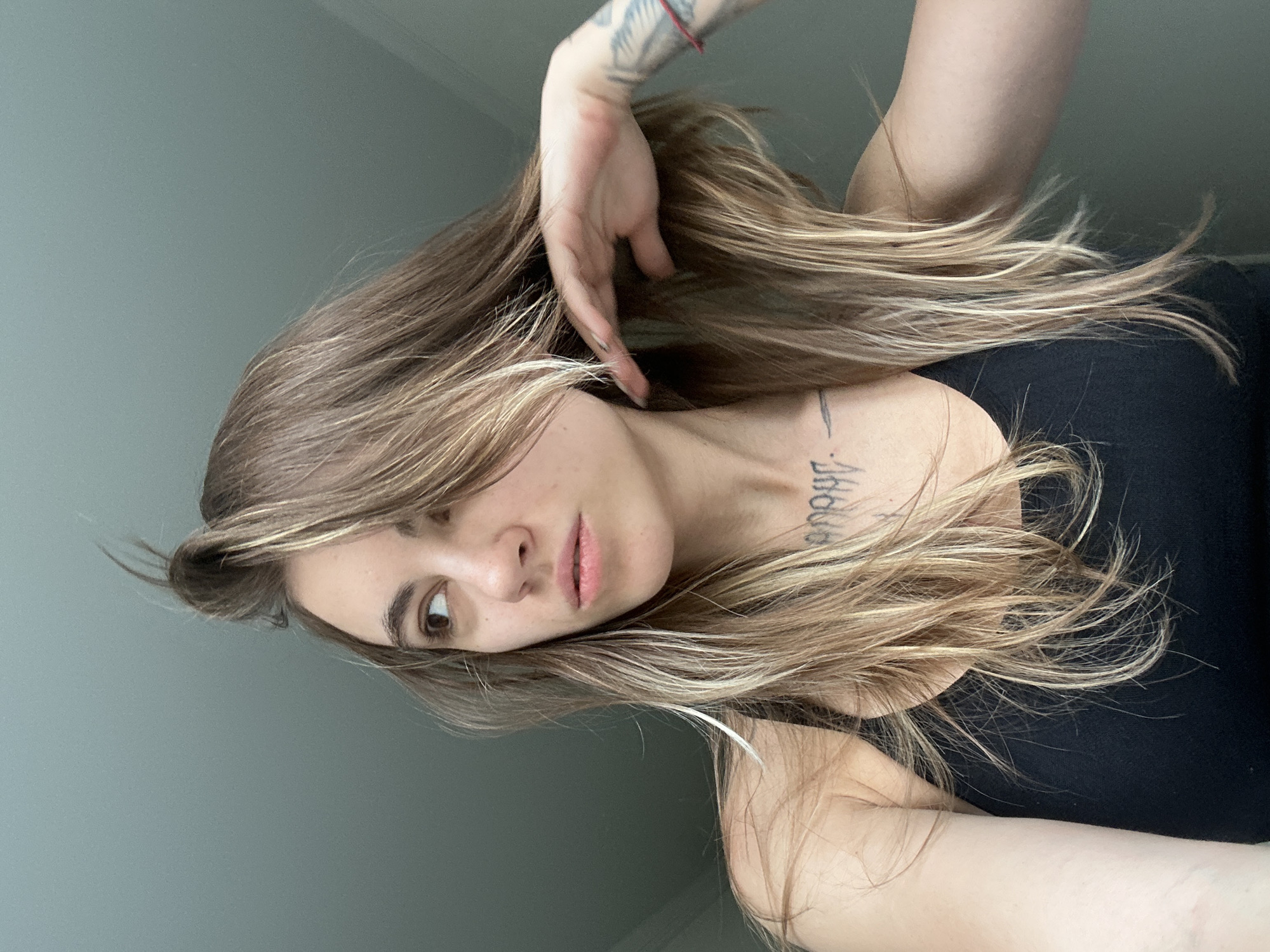 A young woman with long, blonde and brown hair is looking to her left. She is wearing a black tank top and has a tattoo on her chest. Her right hand is raised, touching her hair. The background is a plain, light gray wall. The image appears to be taken indoors with natural light. The location of Moscow, Russia is not present in the image.