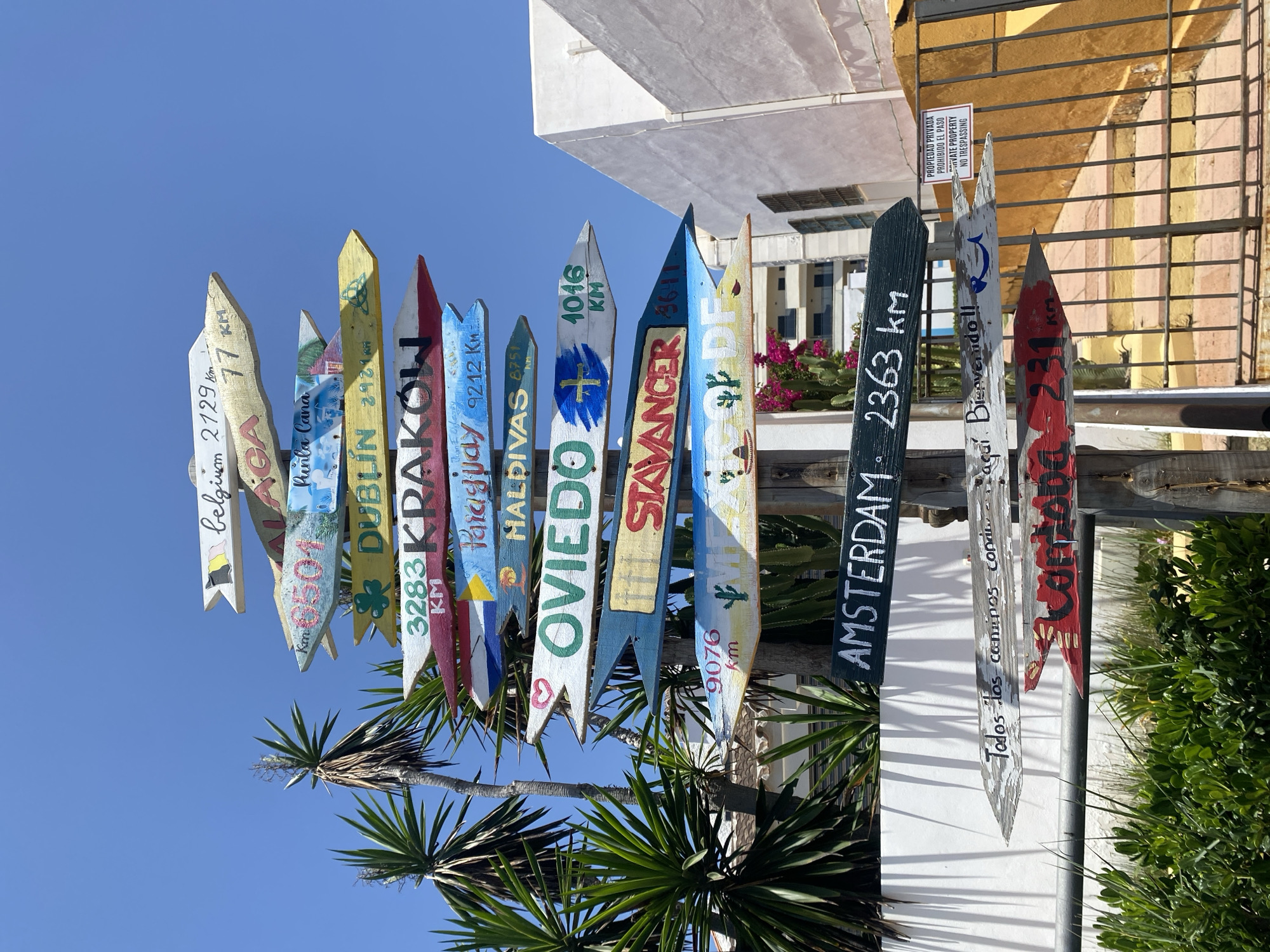 A weathered, multi-directional signpost stands outdoors against a bright blue sky. The signpost is made of wooden arrows, each pointing in a different direction and bearing painted text and distances. The signs list various destinations such as "BELGIUM 2729 km", "MALAGA 77 km", "PUNTA CANA", "DUBLIN 2921 km", "KRAKOW 3283 km", "PARAGUAY 9212 Km", "HALDIVAS 8751 Km", "OVIEDO 1046 KM", "SAVANGER 3611", "AMSTERDAM 2363 km", "Todos los caminos conducen a Roma. Bienvenido !!", and "CORDOBA 231 KM". Some signs have small painted illustrations, like a flag, a shamrock, a stylized wave, a cactus, a sombrero, and a smiling fish. Palm trees, cacti, and green foliage are visible around the base of the signpost, along with a white building and a yellow fence with a "PRIVATE PROPERTY NO TRESPASSING" sign. The scene appears to be in the city of Resinera-voladilla, Spain.