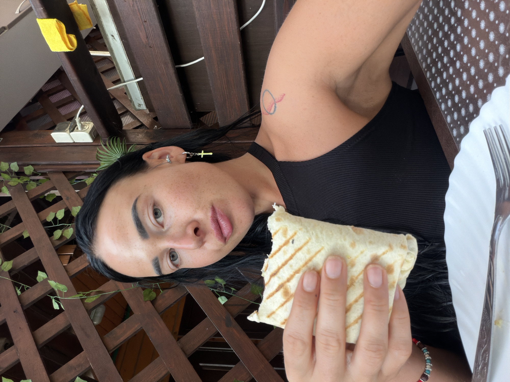 A young woman with black hair and light-colored eyes is holding a partially eaten wrap in her right hand. She is wearing a black tank top, a silver cross earring in her left ear, and a beaded bracelet on her left wrist. There is a small tattoo on her right shoulder. She is seated at a table with a white plate and a fork. The background consists of a wooden lattice structure and some green vines. The setting appears to be outdoors, possibly on a balcony or patio, in Sochi, Russia.