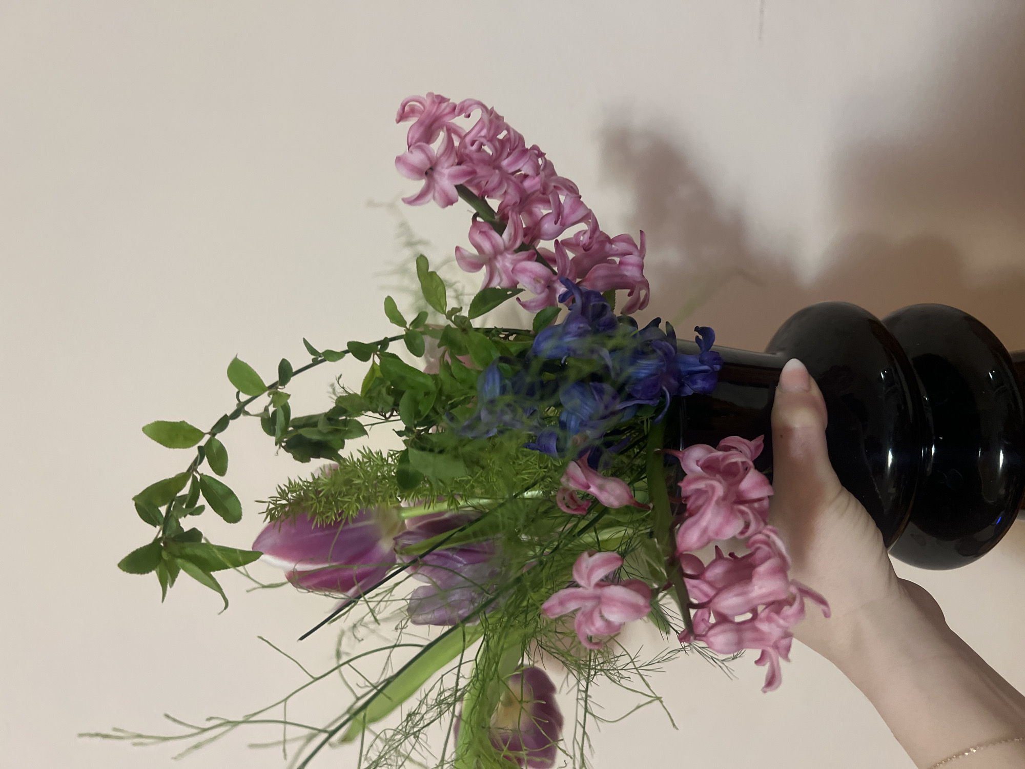 The image shows a floral arrangement in a shiny, dark-colored vase. The arrangement includes pink hyacinths, blue delphiniums, purple tulips, and various green foliage and filler. A light-skinned human hand is holding the vase. The background is a plain, light-colored wall.
There is no information in the image to suggest the location is in the city of Plovdiv, Bulgaria. Therefore, the location is unknown.