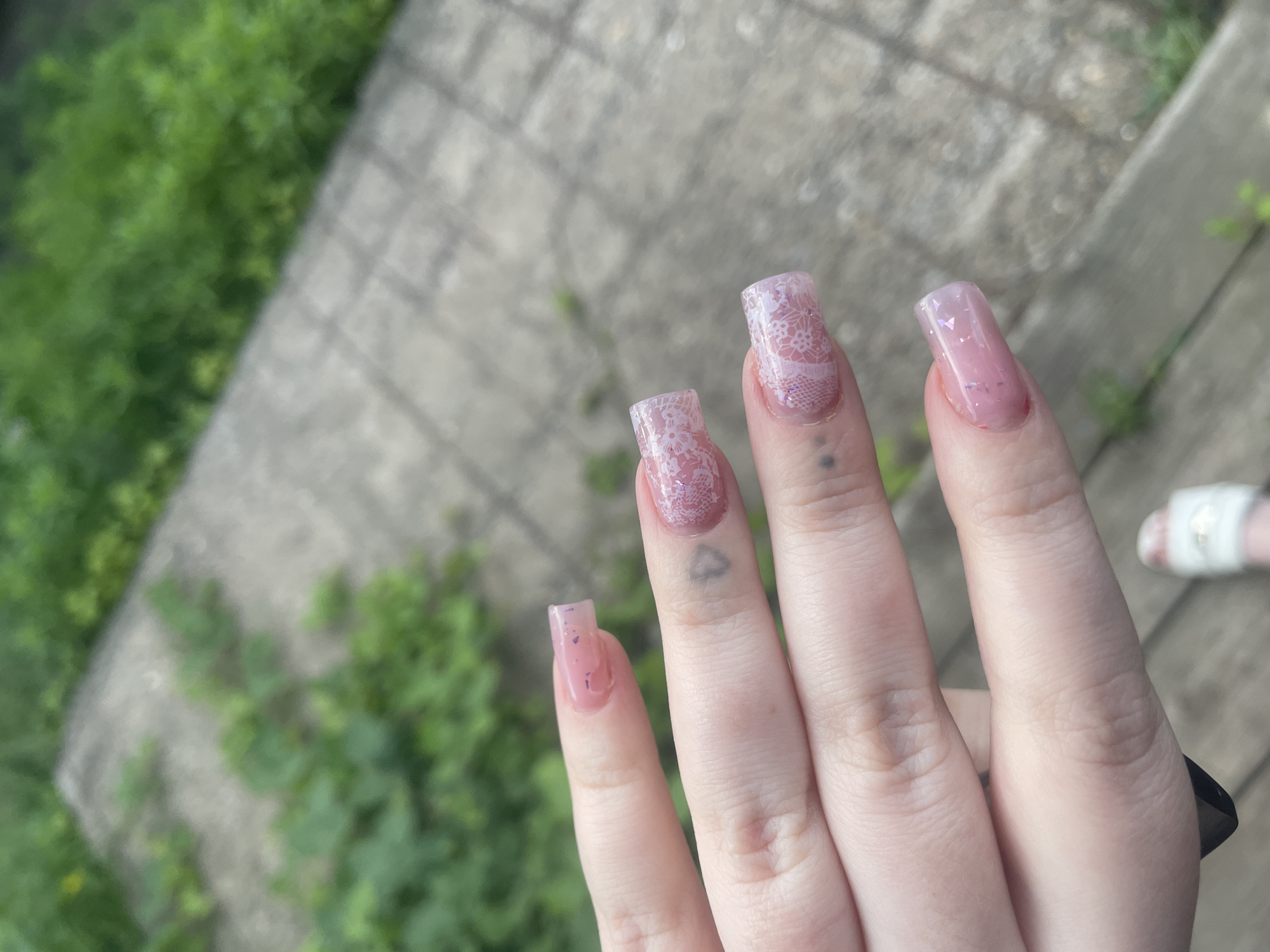 A close-up shot focuses on a person's left hand, adorned with meticulously manicured, square-shaped artificial nails. The nails are a light pink hue, with subtle glitter embedded within the polish. Two of the nails, specifically the ring and middle fingers, feature intricate white lace patterns applied over the pink base. Faint, dark grey heart-shaped tattoos are visible on the skin of these fingers.
The hand is positioned over a grey, paved surface that appears to be outdoors. The paving stones are laid in a grid pattern. Lush green foliage, some of it overgrown and wild, is visible in the background, suggesting a natural or semi-natural outdoor setting. The lighting suggests it is daytime, likely overcast given the soft illumination.
The location of Линёвка, Russia is not discernible from the provided image alone.