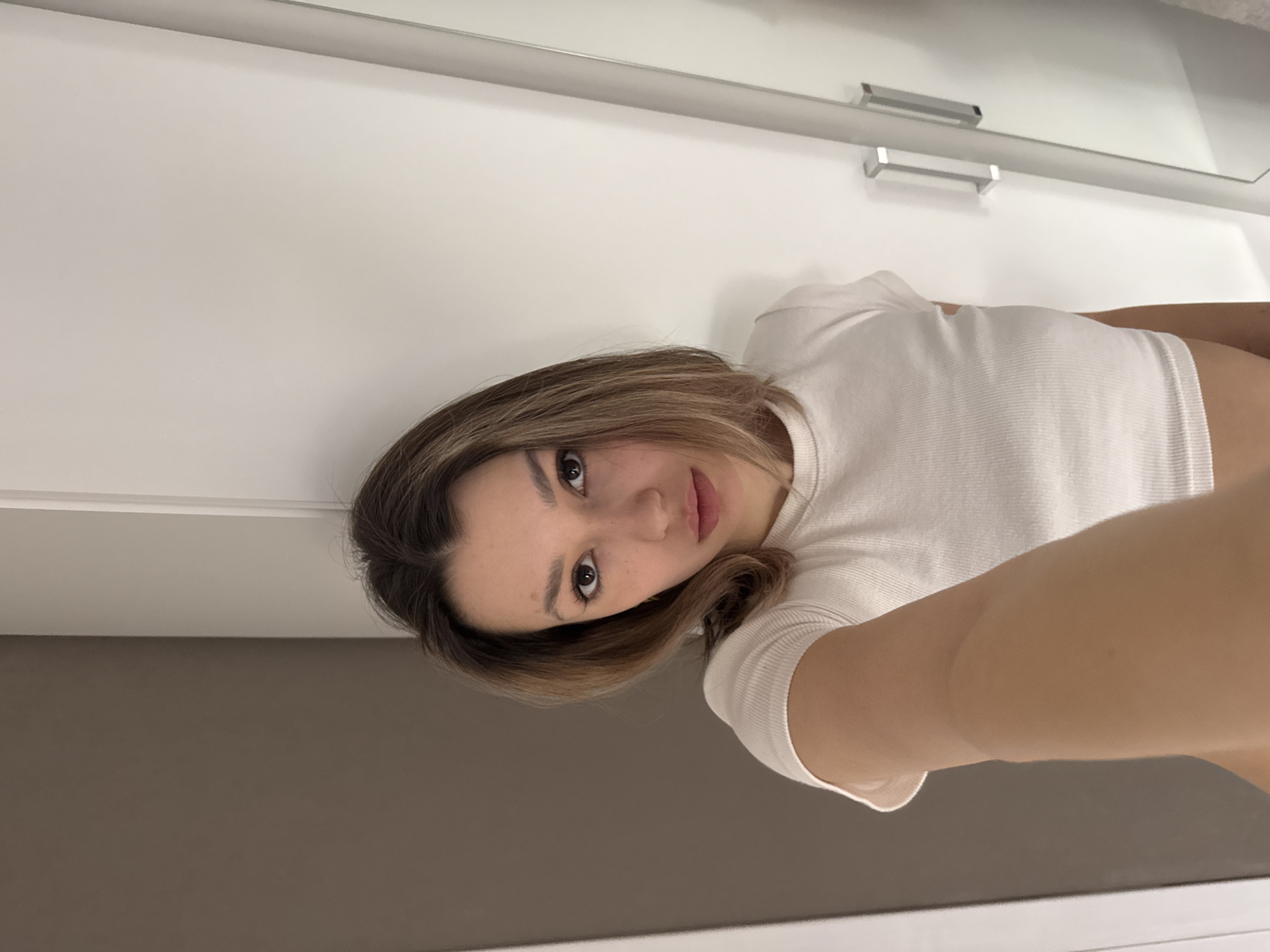 An indoor, vertically oriented selfie captures a young woman looking directly at the camera with a calm, neutral expression. She appears to be in her late teens or early twenties, with medium-length brown hair featuring lighter highlights, framing her face. She has dark brown eyes and a faint mole or freckle near her right eye. She is wearing a white, ribbed, short-sleeved top.
The background consists of a bright white surface, likely a wall or cabinet, adorned with two silver rectangular handles and a horizontal silver bar or shelf. Below this white area, the lower part of the image shows a solid, dark brownish-gray surface, possibly a wall. Her arm is visible, extended as she holds the camera, indicating she is taking the photograph herself. The lighting is bright and evenly diffused, suggesting an interior space, possibly a room with modern, minimalist decor. There is no visible text in the image.