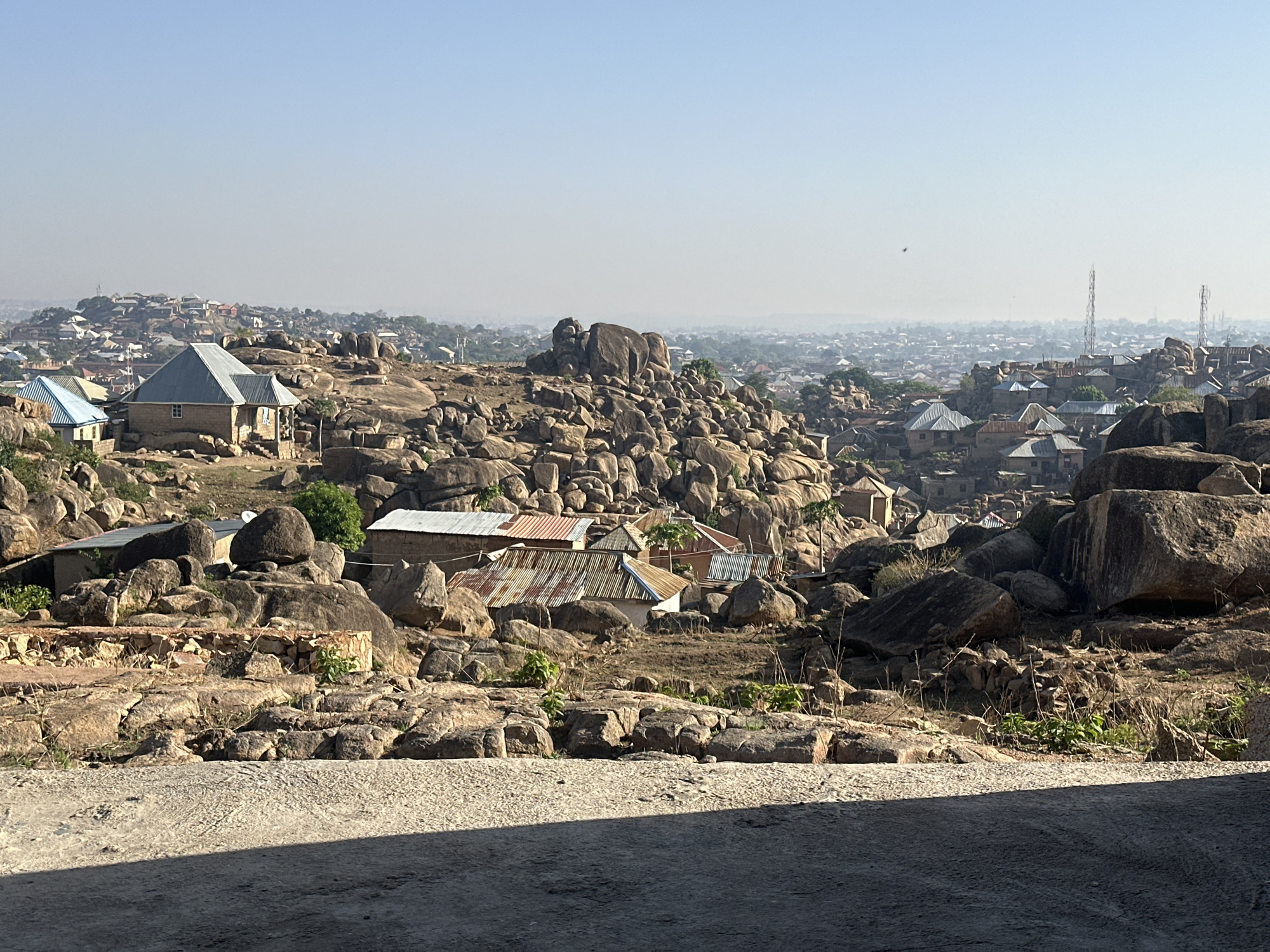 The image shows a landscape of Jos, Nigeria. In the foreground is a rough concrete surface. In the middle ground, a terrain of rocks and boulders dominates the scene, interspersed with sparse vegetation and buildings with corrugated metal roofs. In the background, a hazy cityscape extends into the distance, with faint outlines of buildings and structures. Communication towers are visible on the right.