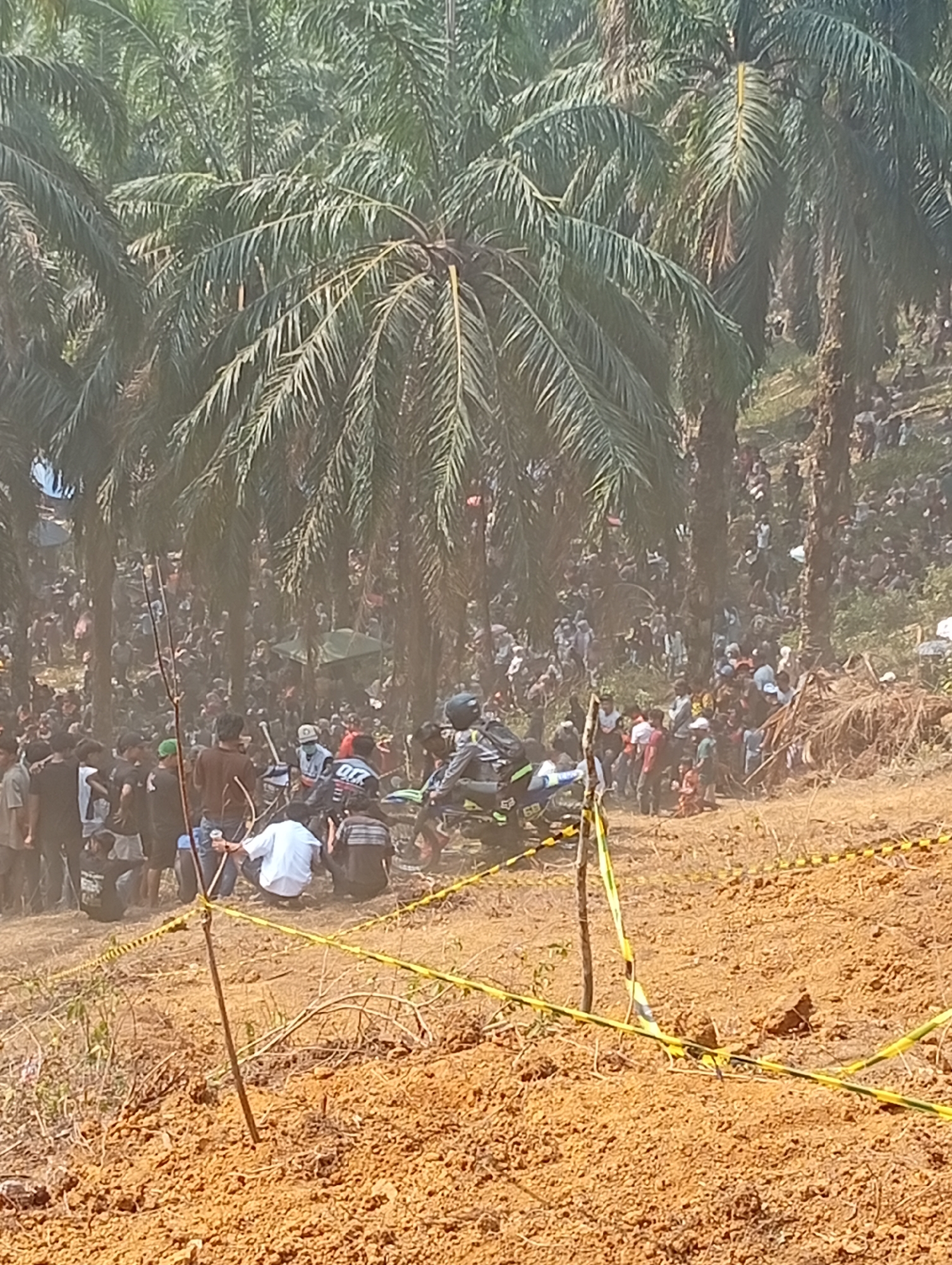The image depicts a motocross event in Sekar Mengkuang, Indonesia, characterized by a large crowd gathered amongst palm trees. In the foreground, a dirt track is visible with yellow and black hazard tape marking the boundaries. Several individuals are present in the foreground, some appearing to be participants with motocross gear and bikes. One motorcycle, predominantly green and white, is positioned to the left of the frame, with a rider wearing a helmet. A single wooden stake with hazard tape is planted in the dirt.
Further into the scene, a dense crowd of spectators is situated on a hillside interspersed with numerous palm trees. The crowd appears to be observing the event, with individuals in various casual attire. Some are standing, while others are seated or crouching. The atmosphere is dusty, suggesting activity from the racing or the dry conditions. In the background, more palm trees extend up the hillside, and hints of tents or canopies are visible amongst the trees. The overall impression is of a lively outdoor event with a significant turnout in a tropical environment.
